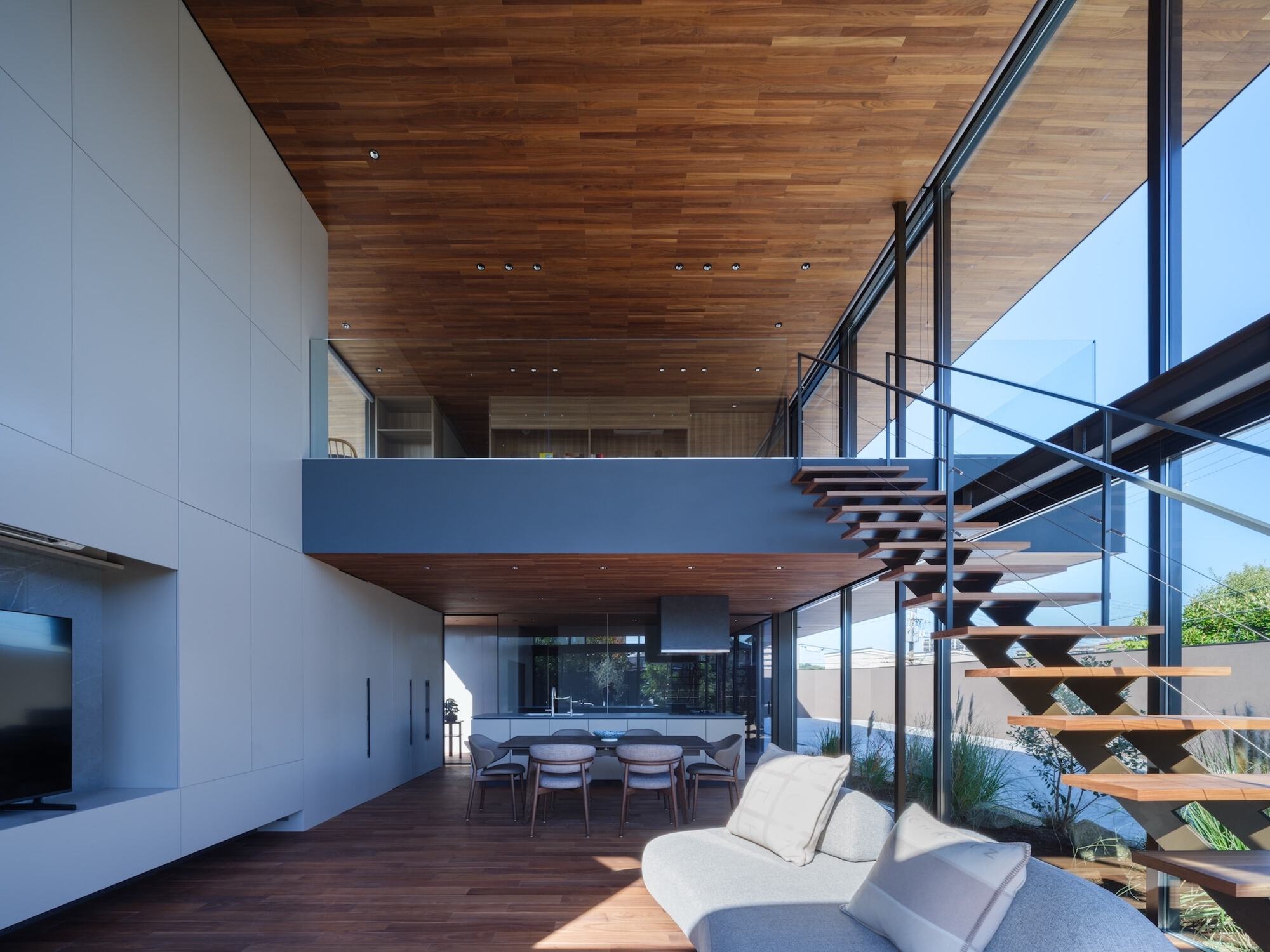 Double-height living space with open-tread timber stair, dining table below a mezzanine and a grey sofa in afternoon sunlight