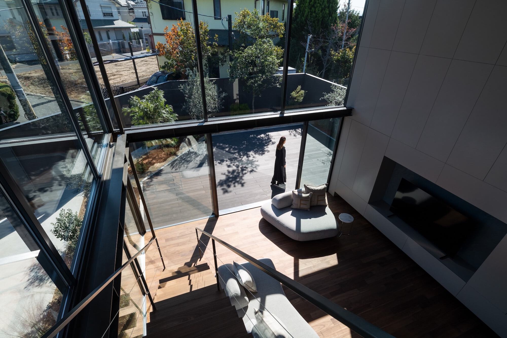 High view down the stair into the living room, where a woman stands on the sunlit deck beyond a curved sofa