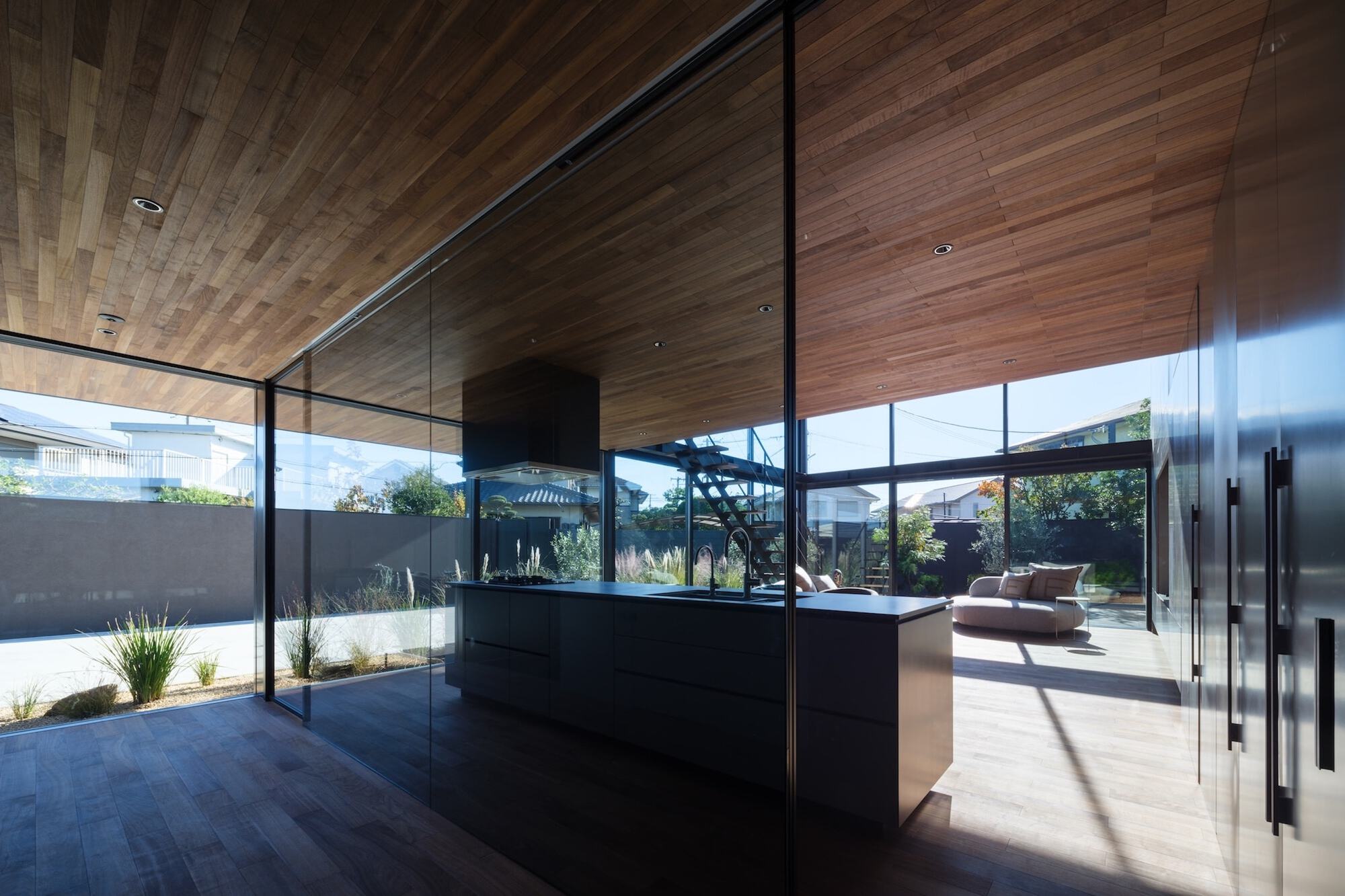 Glass-walled kitchen island beneath a timber slat ceiling, with a sunlit terrace, lounge seating and grasses visible beyond in clear daylight