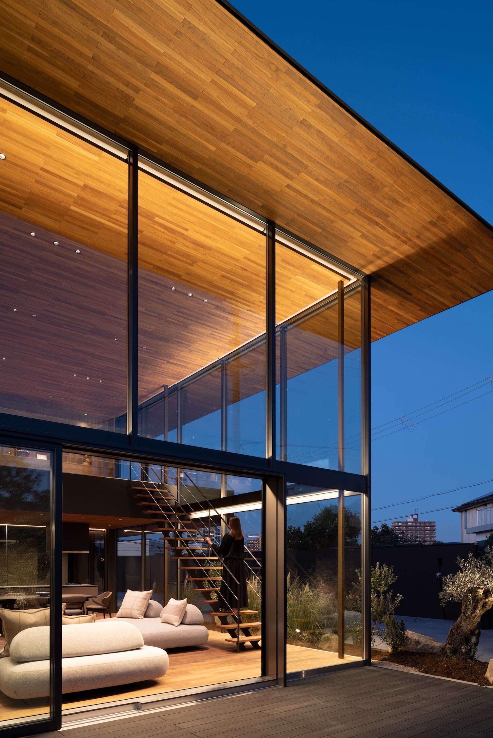 Corner of the glazed living space with an open timber stair, a person standing on it, and the roof soffit glowing at dusk