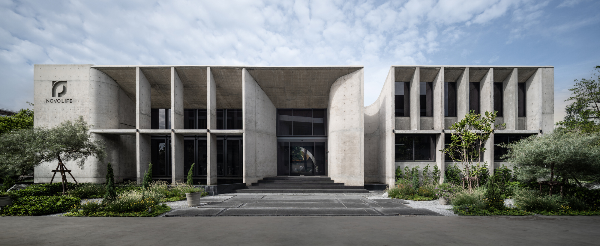 Straight-on elevation of the two-part concrete facade with a central entrance stair and planted beds in soft daylight