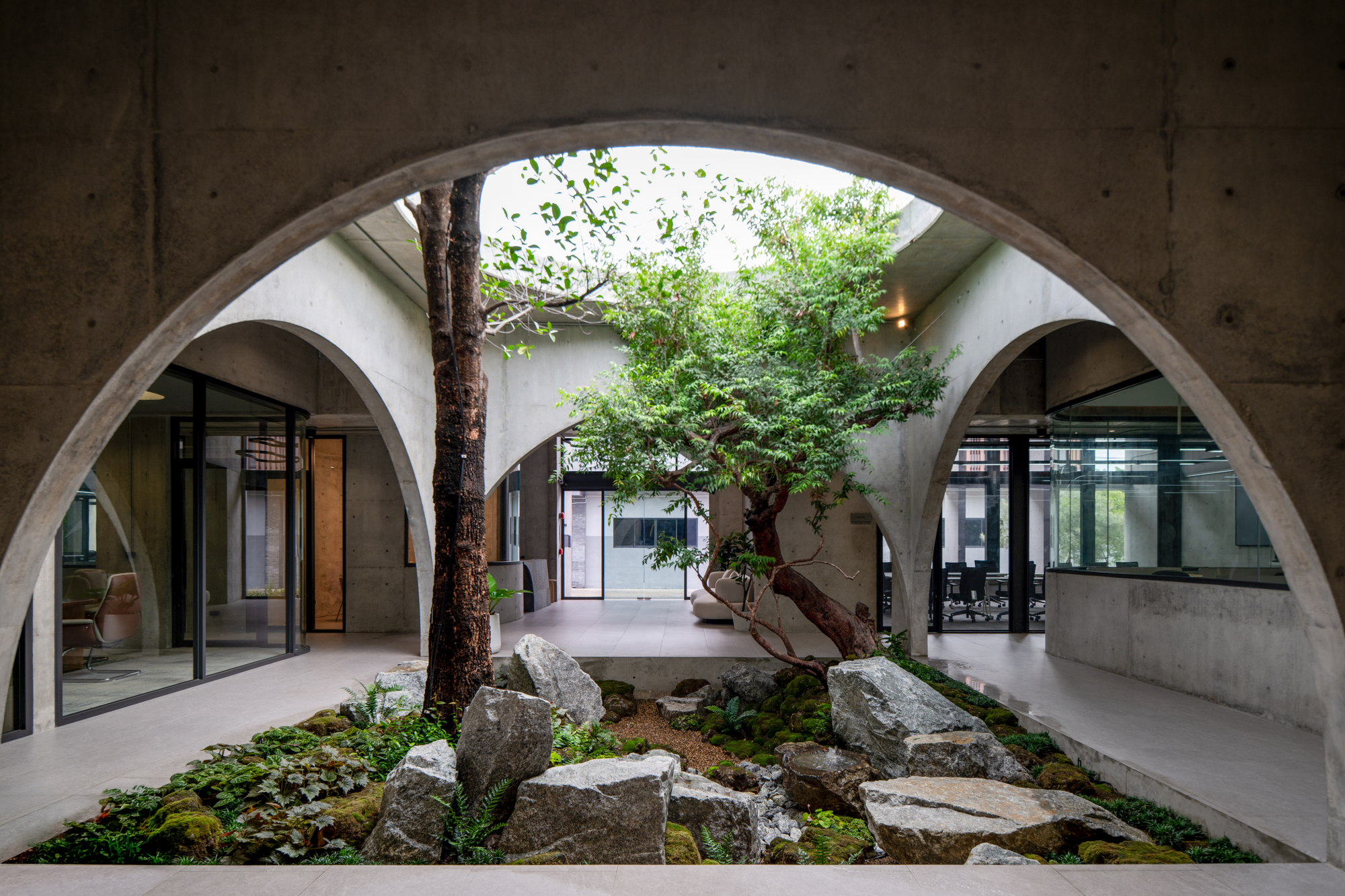 Looking through concrete arches into a courtyard garden of boulders, moss and a leaning tree, with glazed offices on both sides