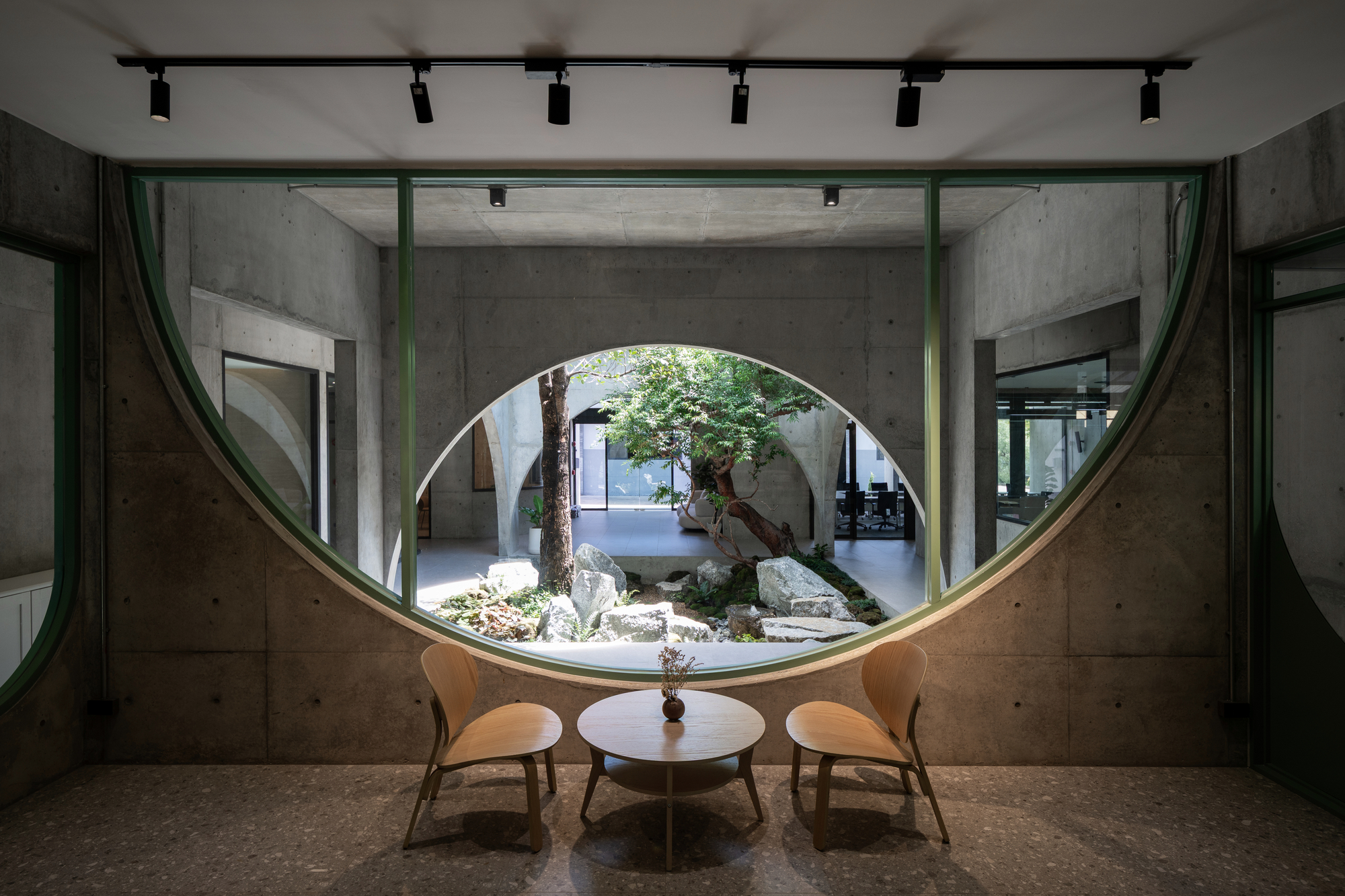 Round green-framed window in a concrete wall, with two wooden chairs and a small table facing the rock garden courtyard