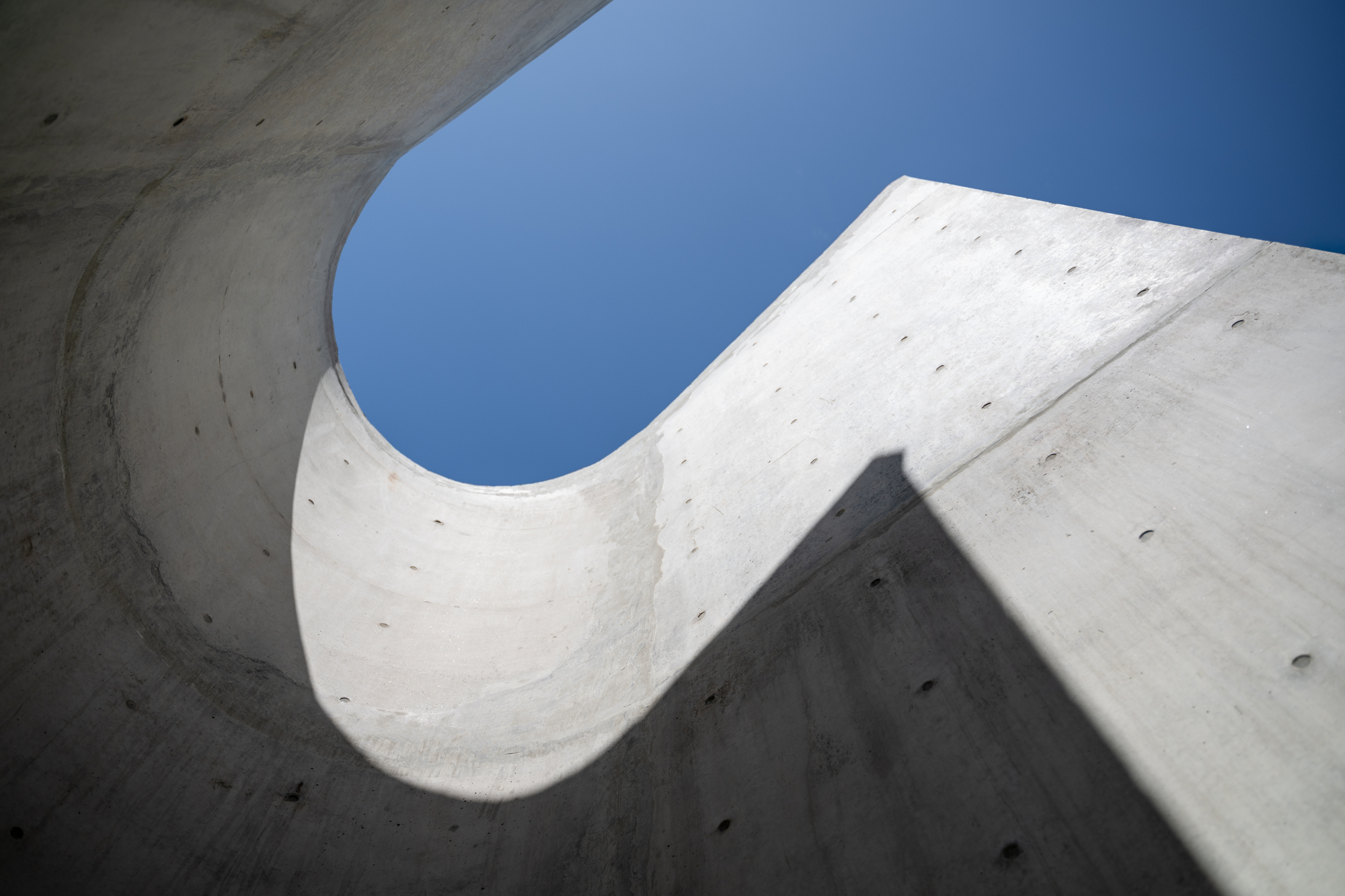 Upward view of a curved concrete wall opening onto blue sky, with a sharp diagonal shadow across the surface