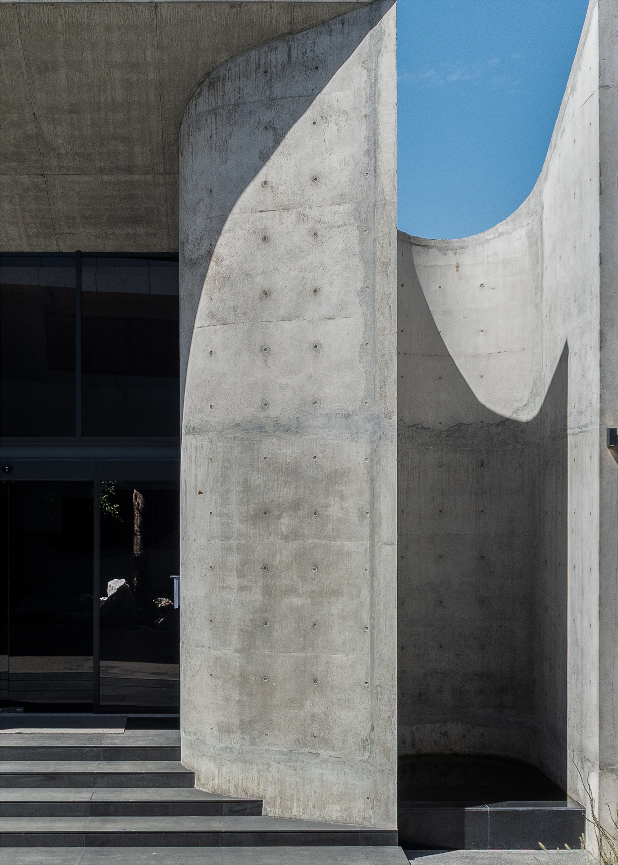 Curved concrete entrance walls with tie-hole patterns beside low black steps, with sunlight casting a diagonal shadow across the surface