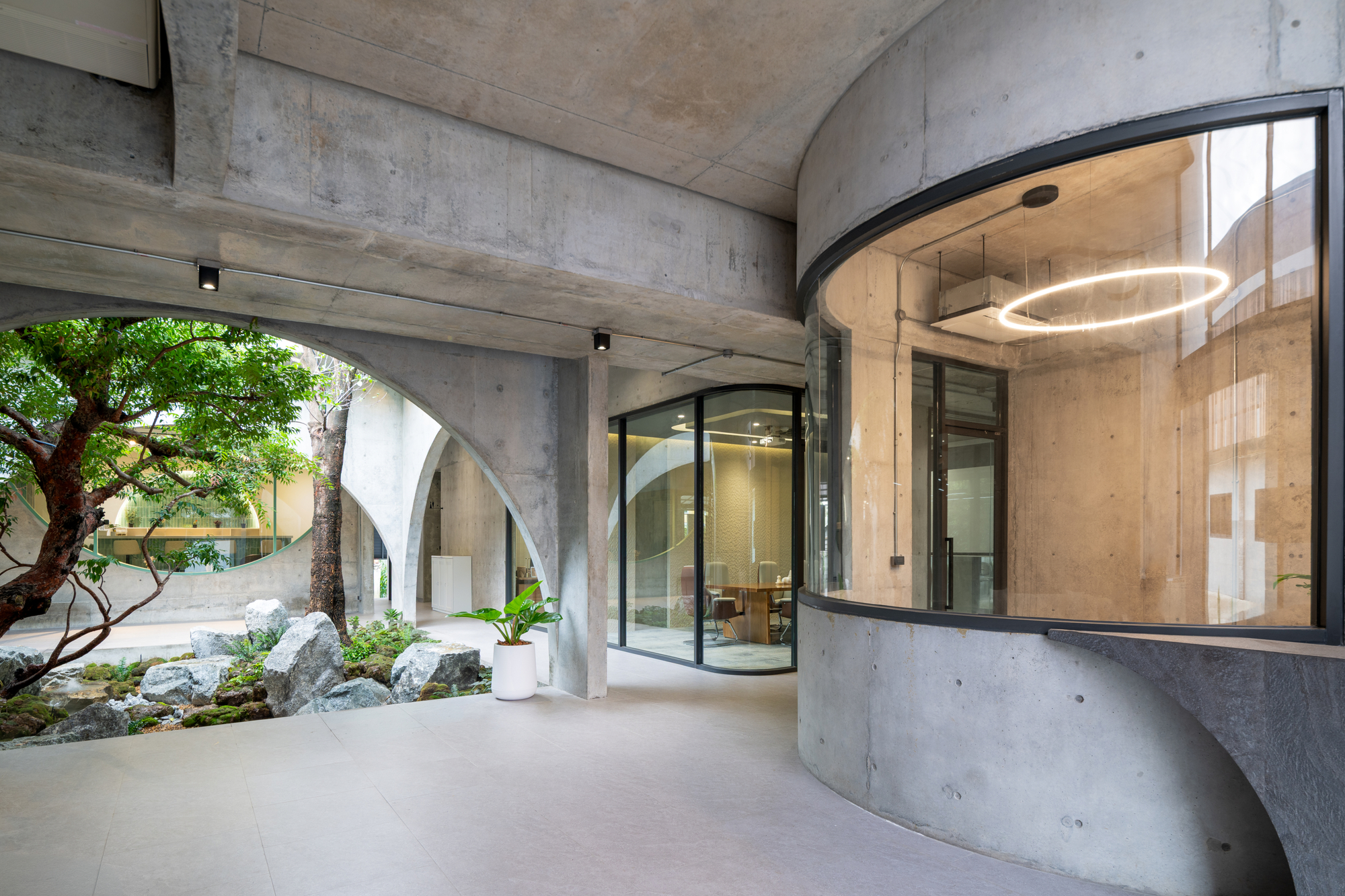 Covered concrete walkway with arched openings onto a rock garden and tree, beside a curved glass meeting room with a ring light
