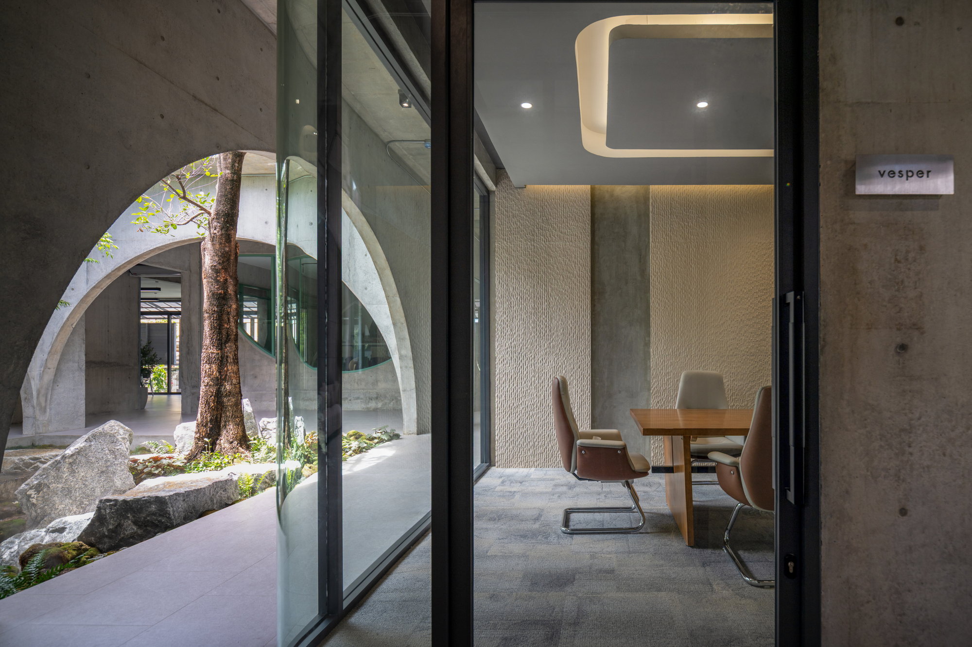 Glass door of a meeting room with textured wall panels, looking back to a courtyard tree framed by concrete arches