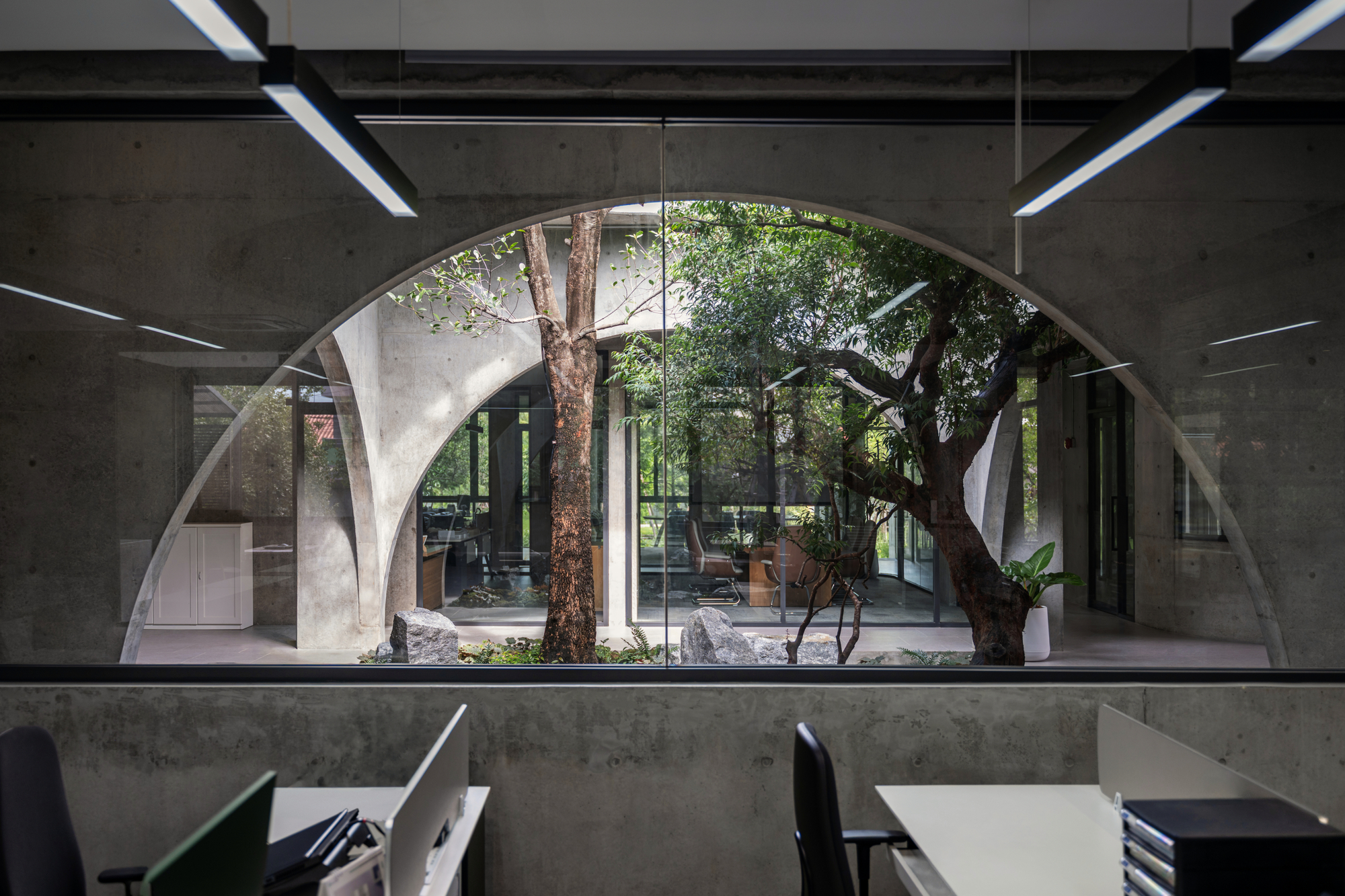 Semicircular arched opening in a concrete wall from the office floor, framing courtyard trees, rocks and glazed rooms beyond