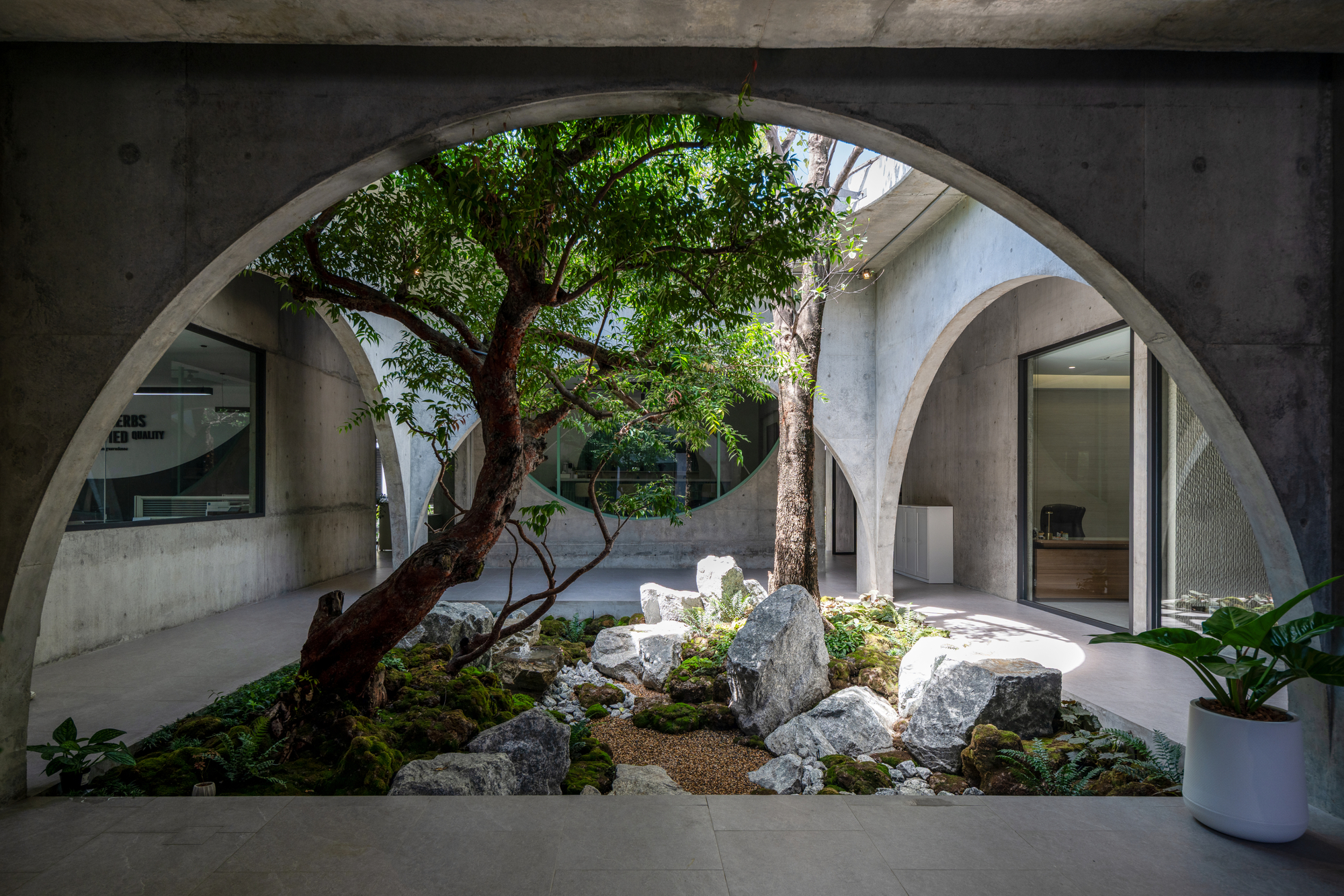 Large concrete arch framing a courtyard garden of mossy boulders, gravel and a leaning tree, with a potted plant in the foreground
