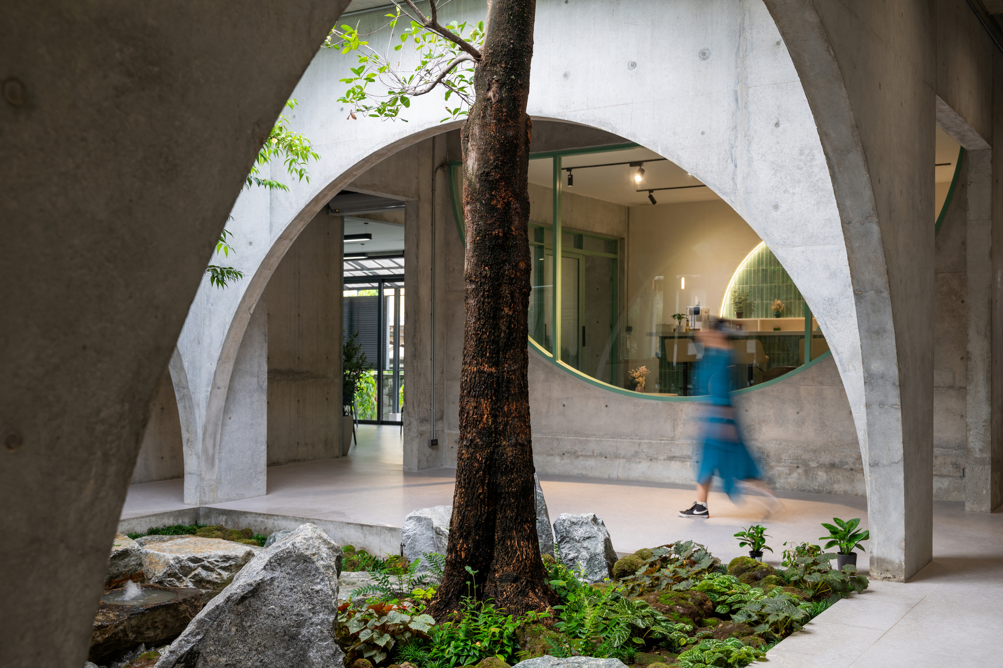 Concrete arches framing a courtyard tree and mossy boulders, with a blurred figure in blue walking past a round window
