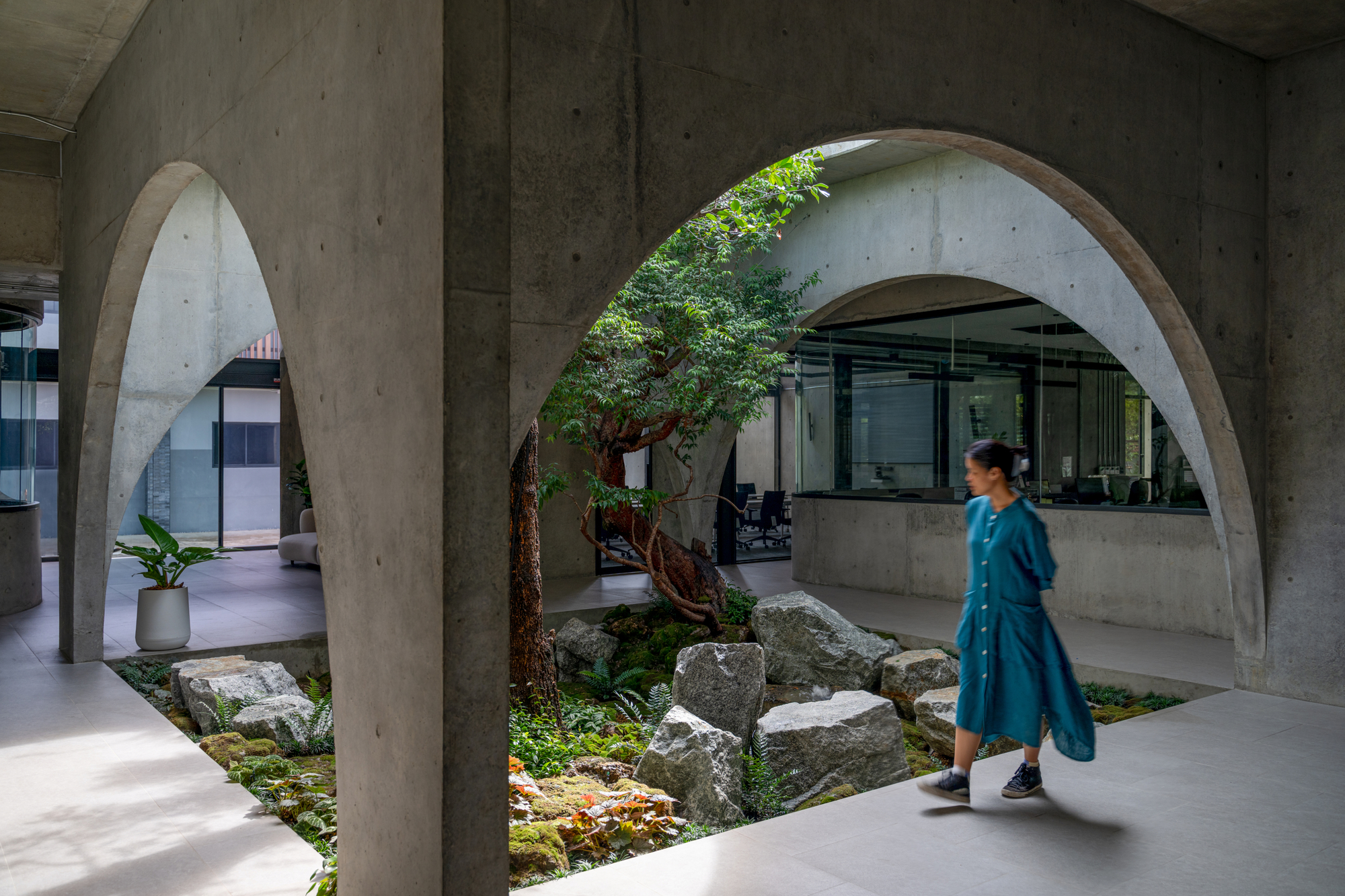 Woman in a teal dress walking beneath board-marked concrete arches beside a courtyard of boulders, moss and a twisted tree