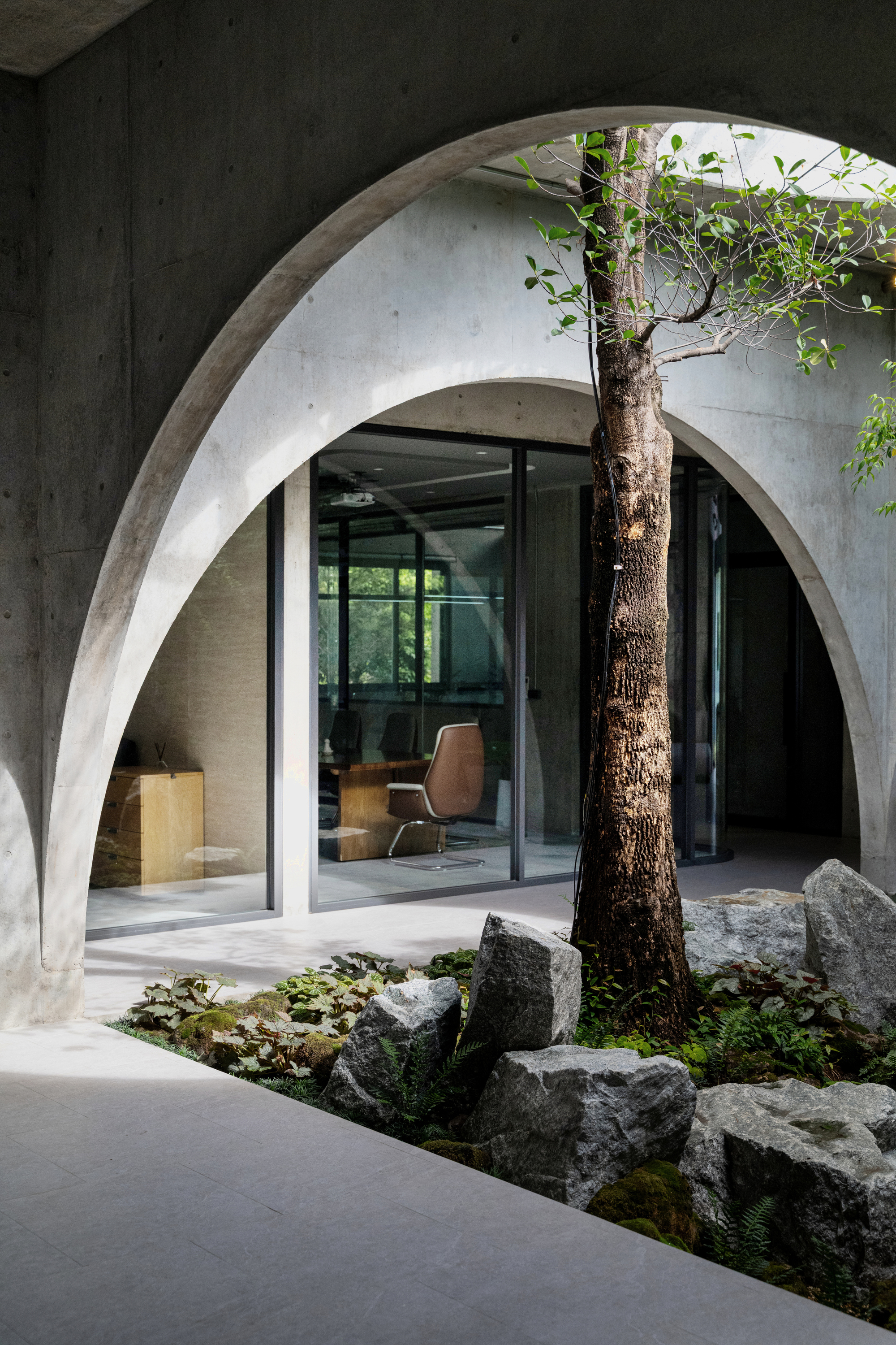 Tall concrete arch framing a courtyard tree and rocks, with a glazed office and leather chair behind in soft daylight