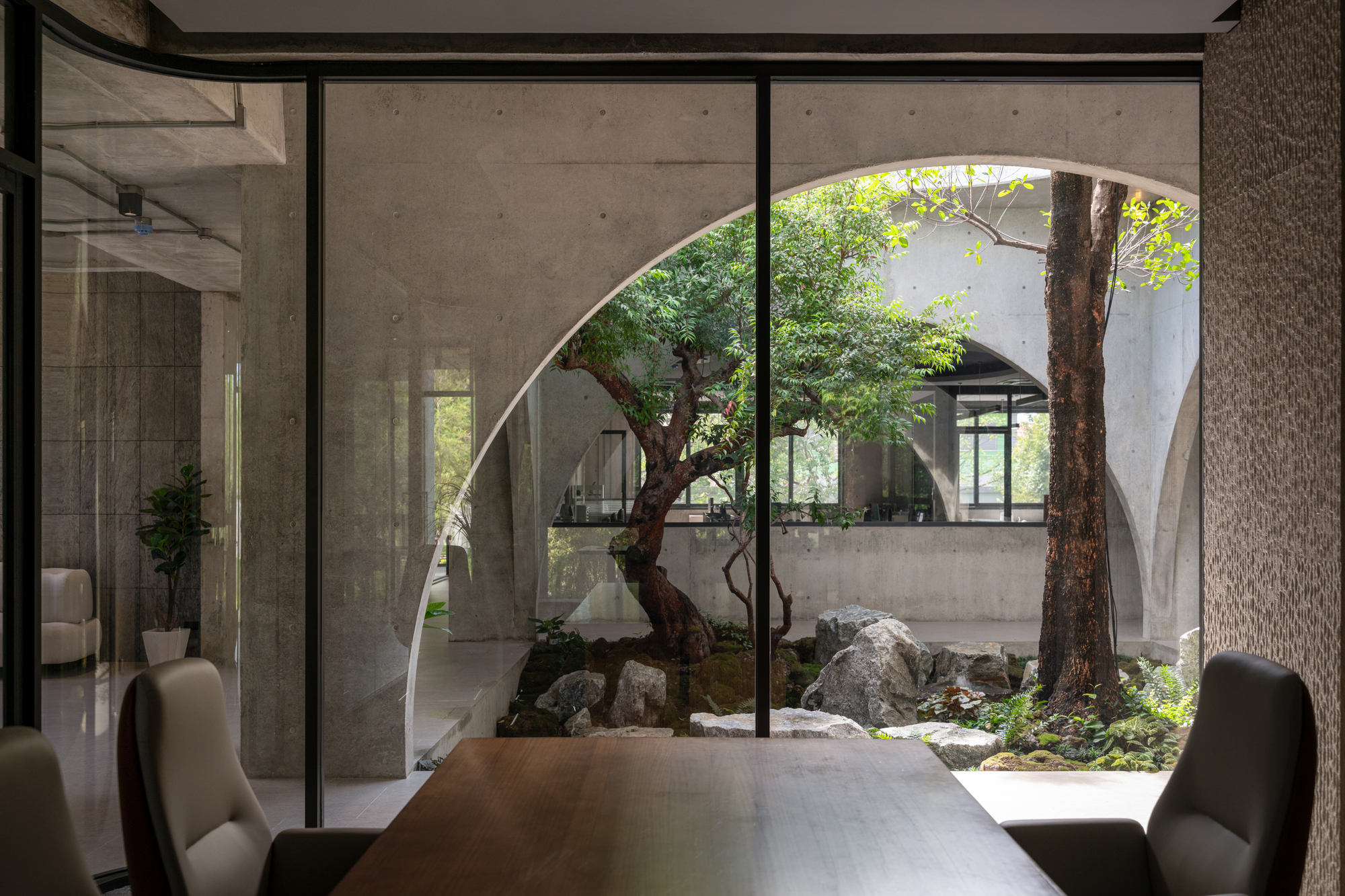 View from a meeting room across a timber table through glass to a courtyard tree, rocks and concrete arches