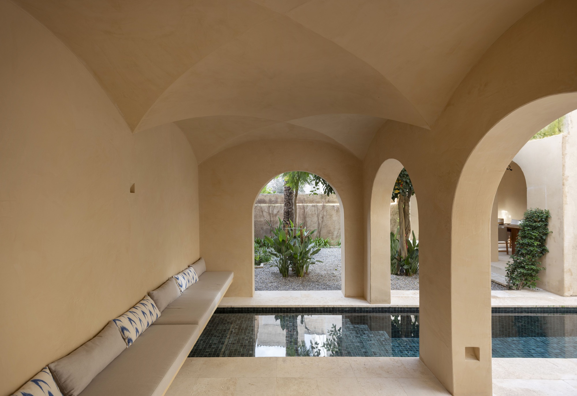 Groin-vaulted loggia with a built-in cushioned bench, arched openings and a tiled plunge pool facing a gravel garden in soft daylight