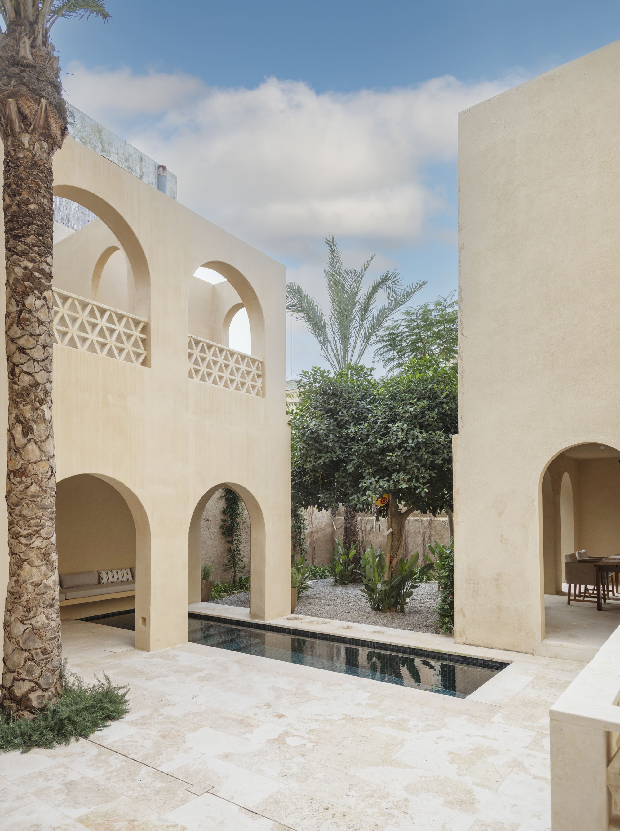 Courtyard of plastered arched walls with lattice balustrades, a narrow pool and an orange tree under a partly cloudy sky