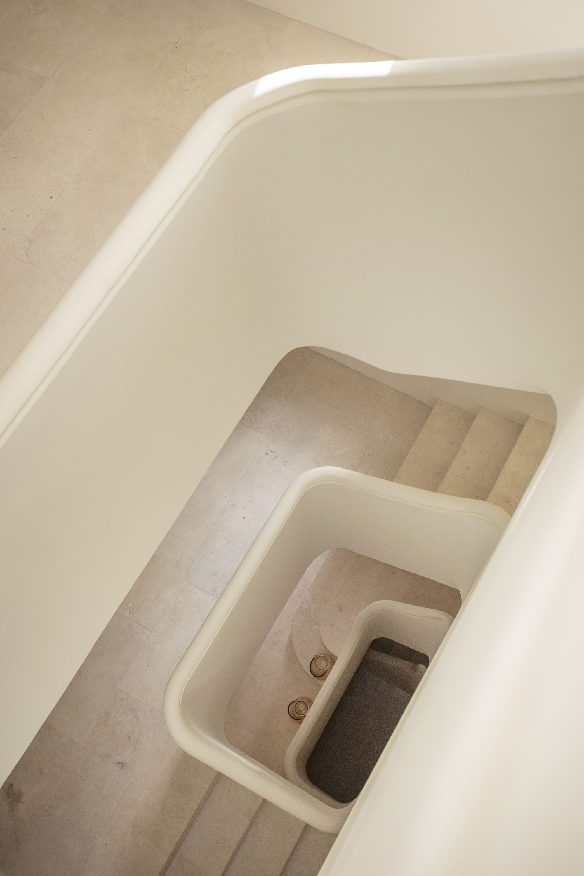 Looking down a curving white stair balustrade onto limestone treads, with soft light grazing the handrail