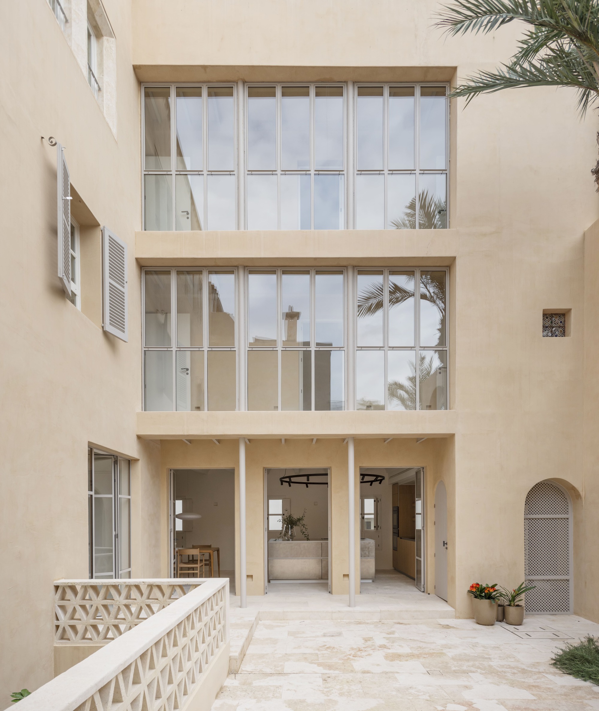 Courtyard facade with two tiers of tall steel-framed windows above open doors, a lattice screen wall and a palm frond overhead