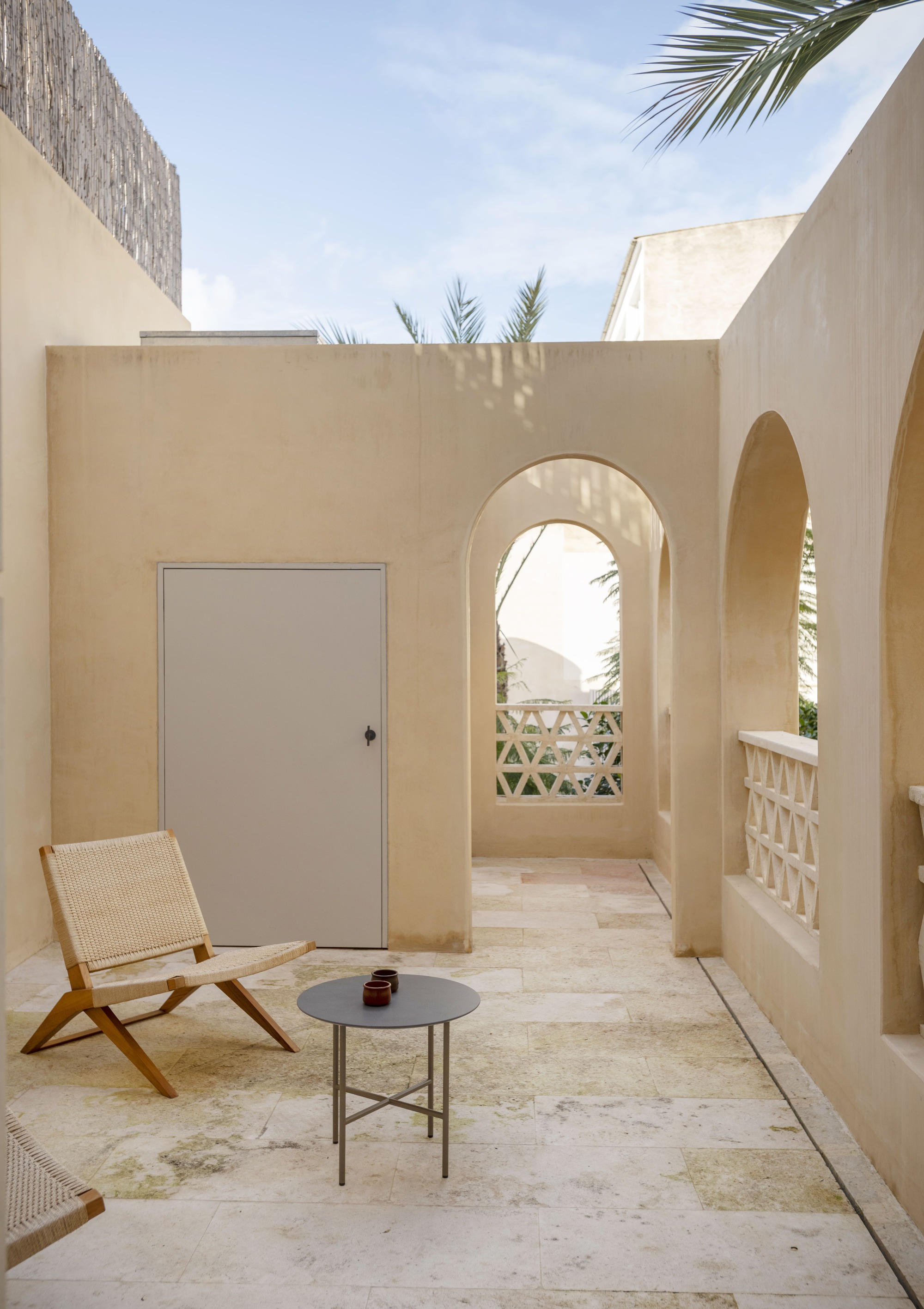 Small terrace with a woven folding chair and round side table beside arched openings and a lattice parapet in afternoon sun