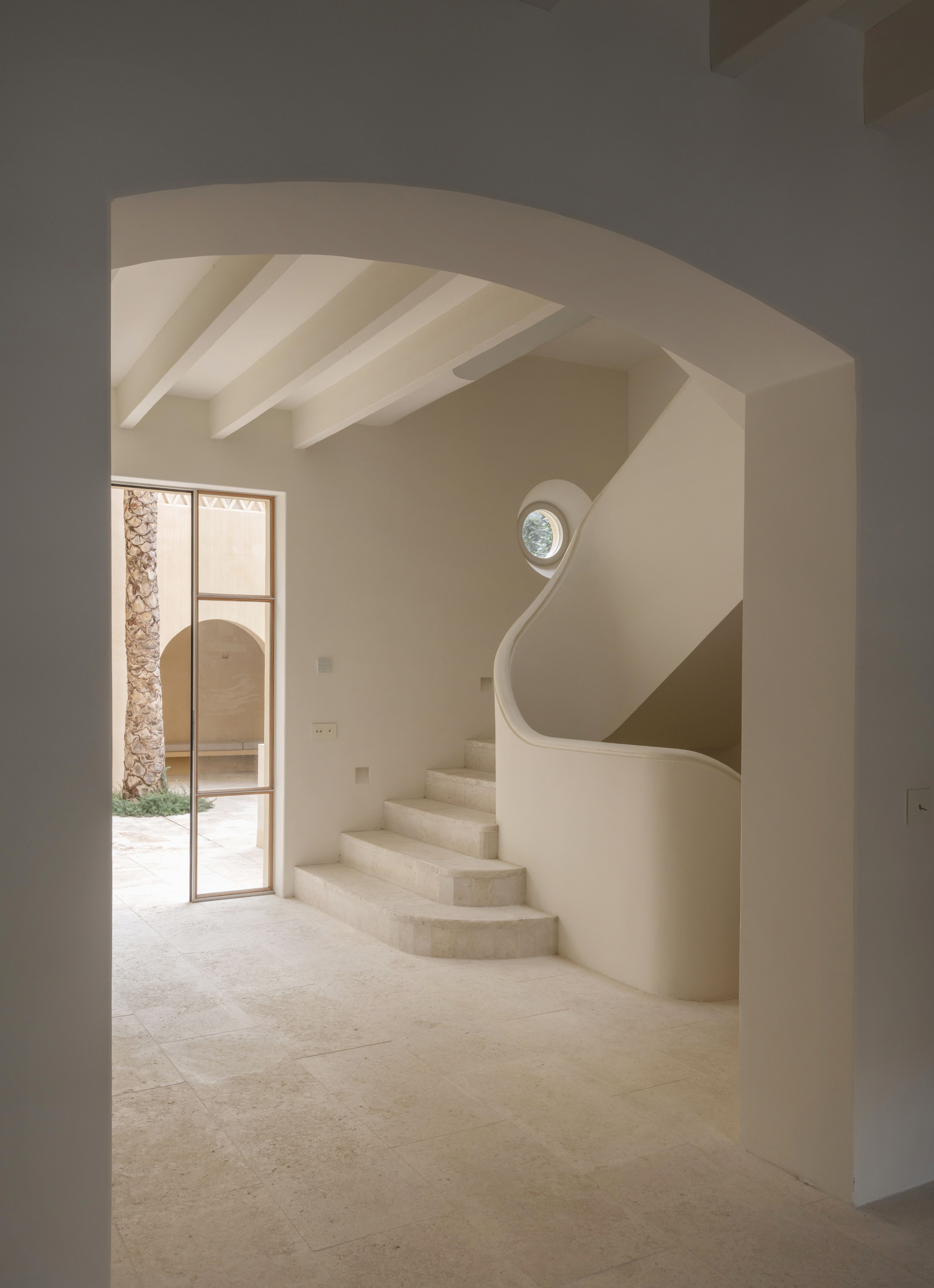 View through a plastered arch to a sculpted curving stair, round window and exposed white beams, with a glazed door open to the courtyard