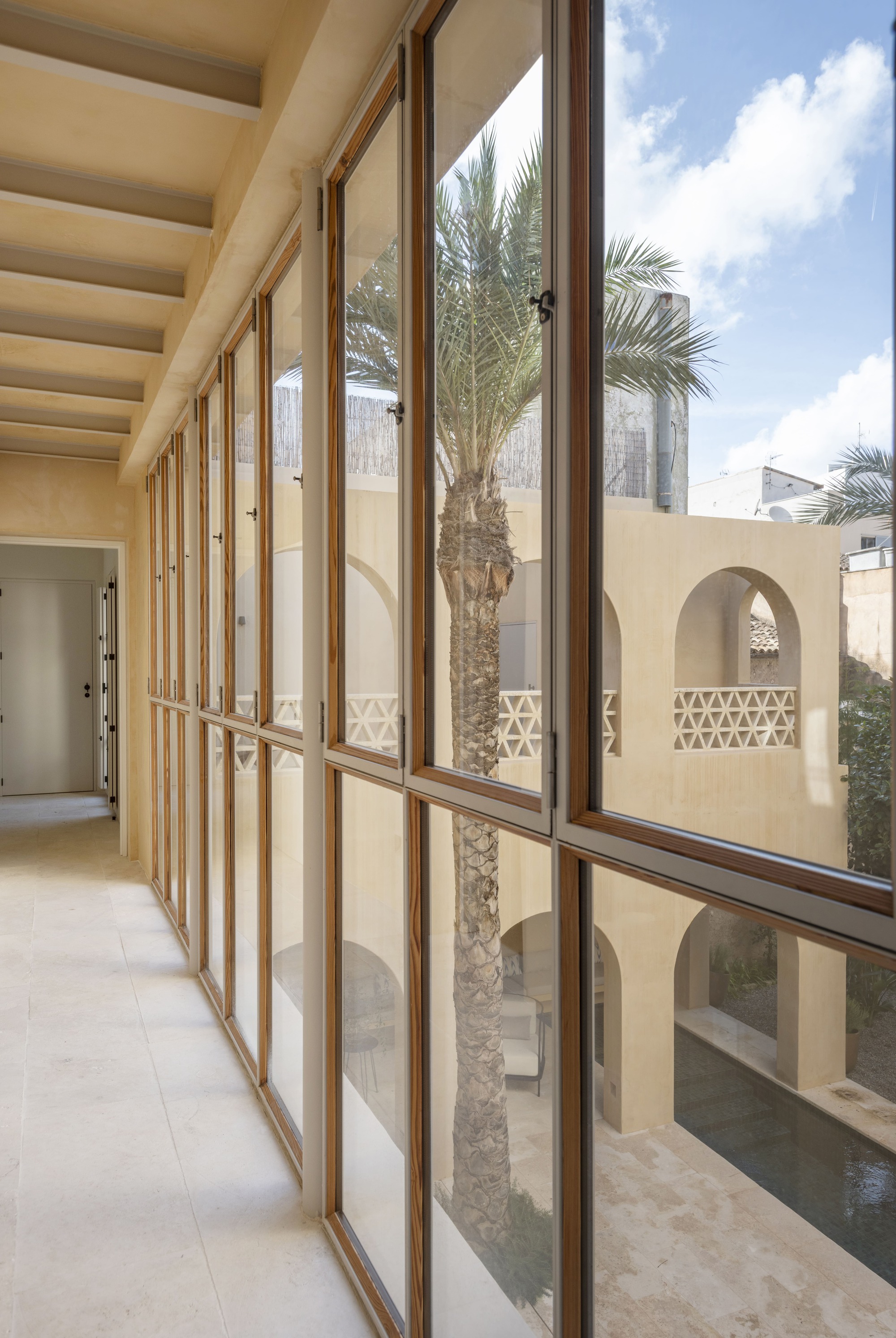 Glazed gallery with timber-framed windows framing a palm tree and arched courtyard walls beside a pool under scattered clouds