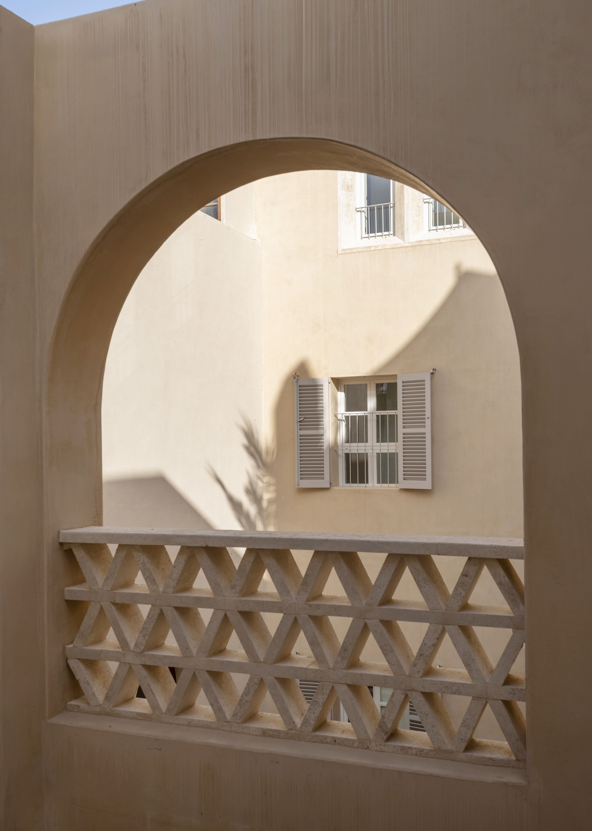 Arched opening in a sand-colored wall with a lattice balustrade, looking onto a shuttered window in afternoon shadow