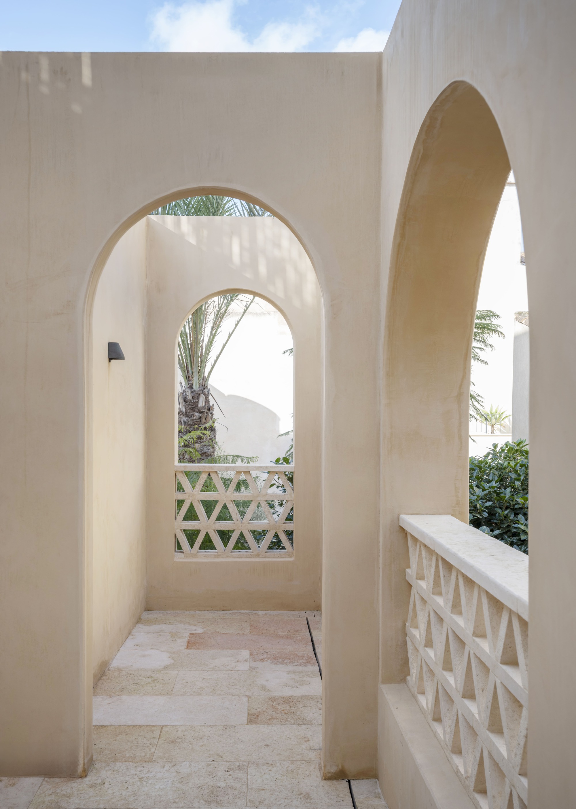 Sequence of rendered arches with triangular lattice screens and a stone-paved terrace, palm fronds visible against a bright sky