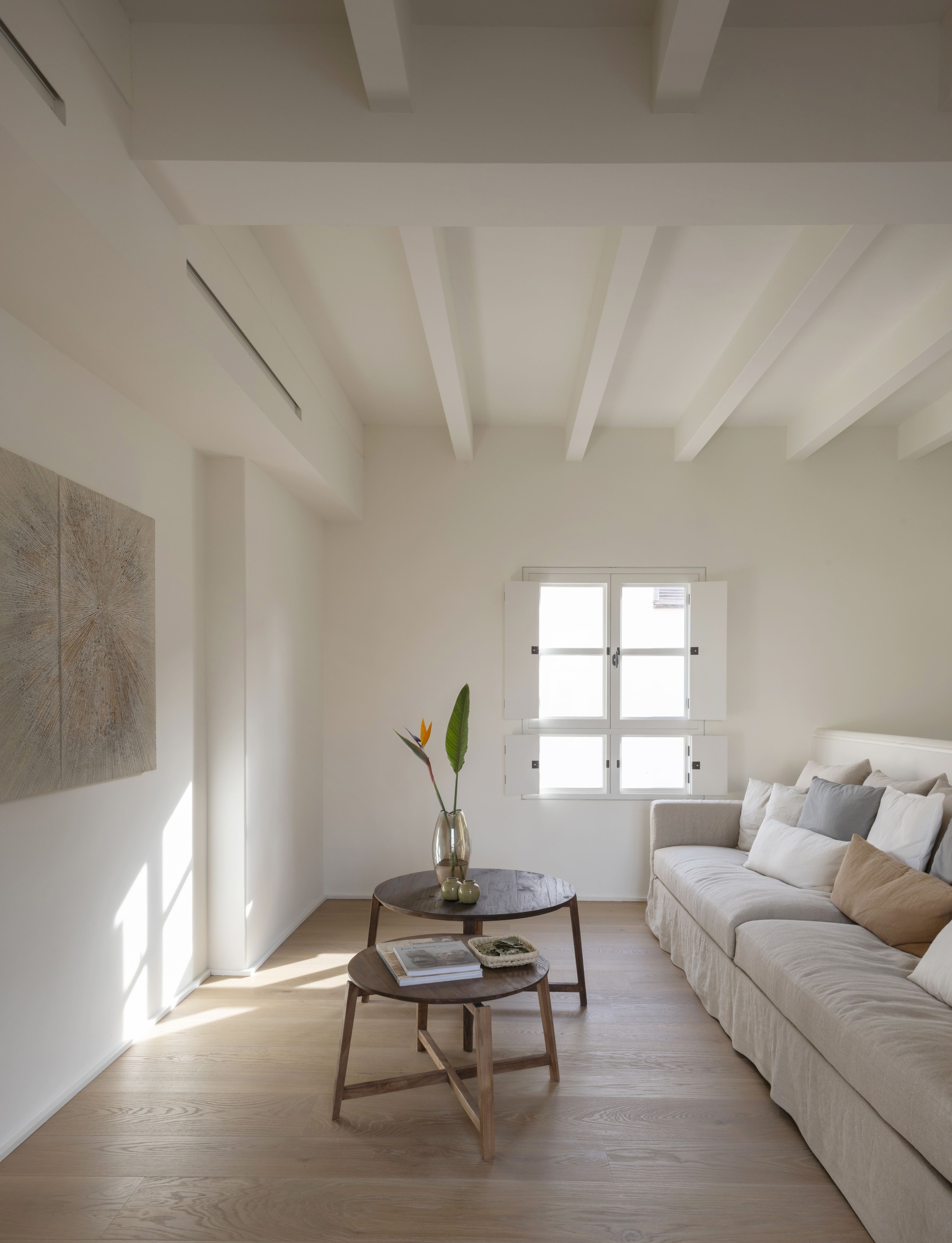 Living room with white painted ceiling beams, a linen sofa and two round timber tables, sunlight falling across the wall