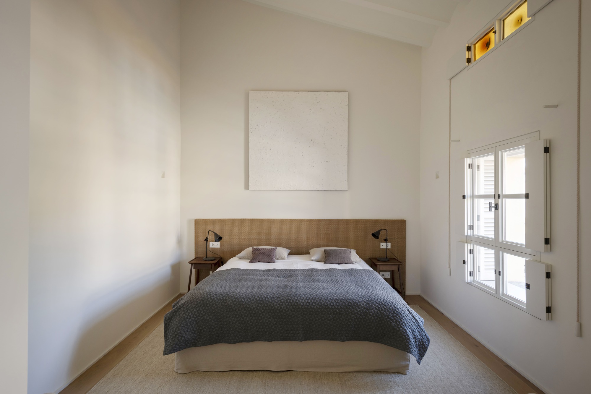 Bedroom with woven headboard, grey quilted bedspread and a tall window with folding shutters in soft daylight