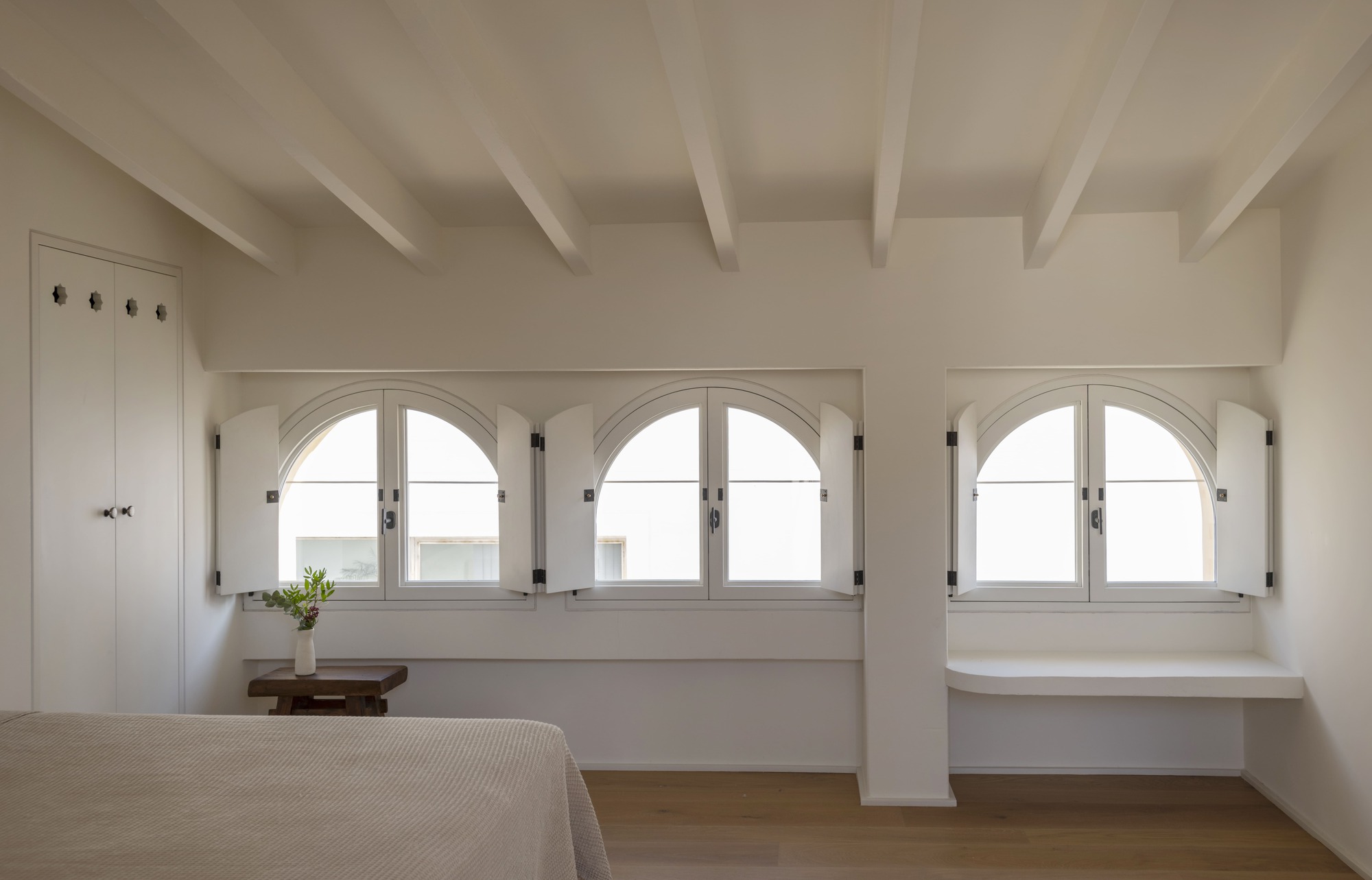Three arched windows with open white shutters beneath exposed ceiling beams, with a built-in shelf and a small bedside table with flowers