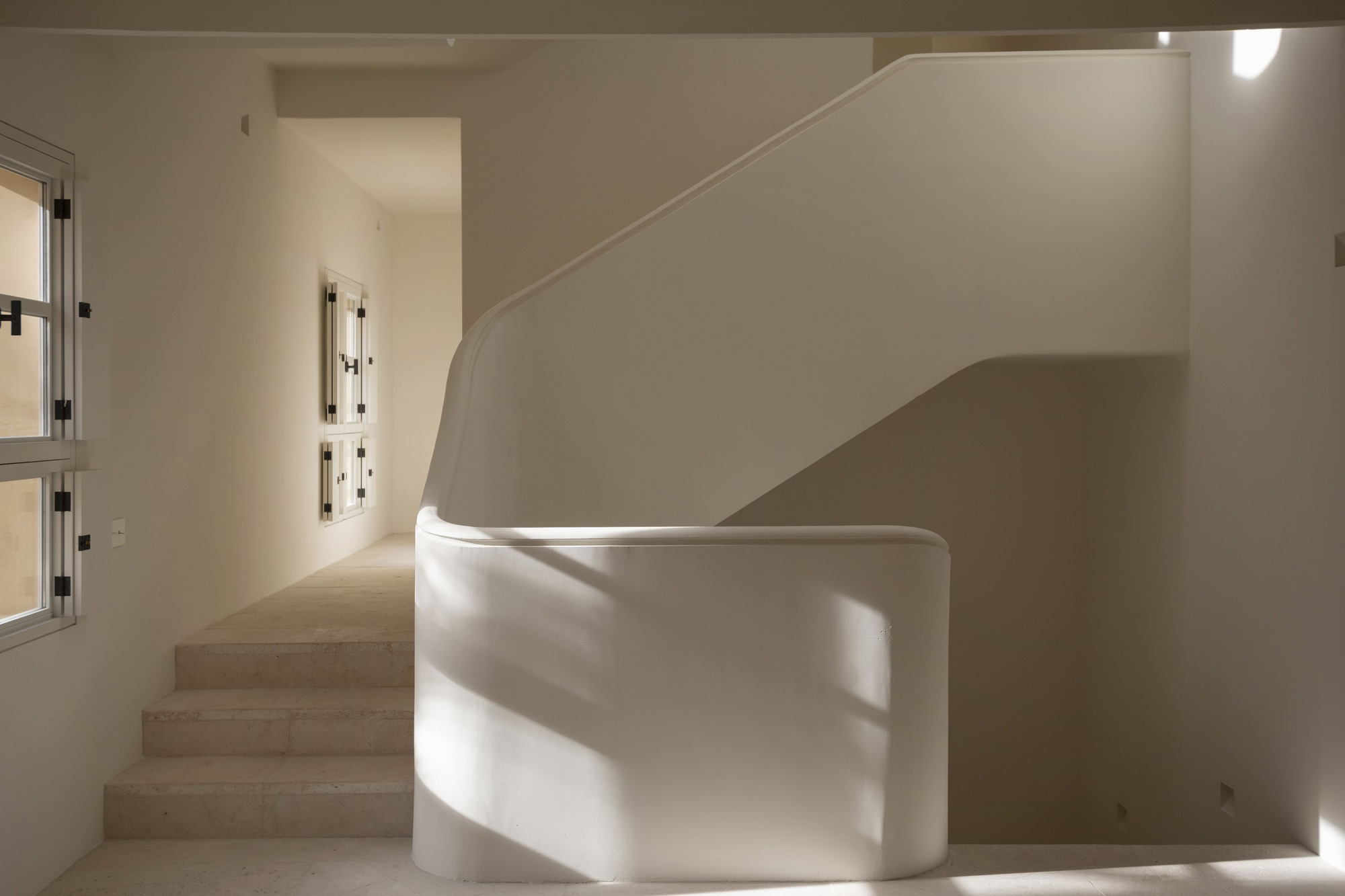 Curved plaster stair balustrade beside stone steps, with sunlight casting soft shadows across the white walls