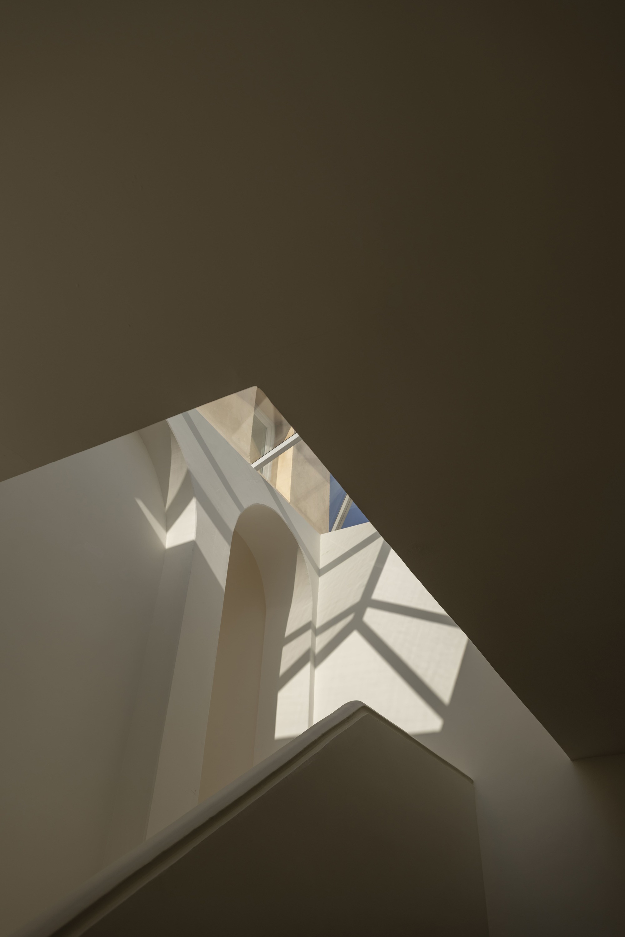 Upward view of a stairwell with an arched recess and a skylight throwing geometric shadows onto the plaster walls