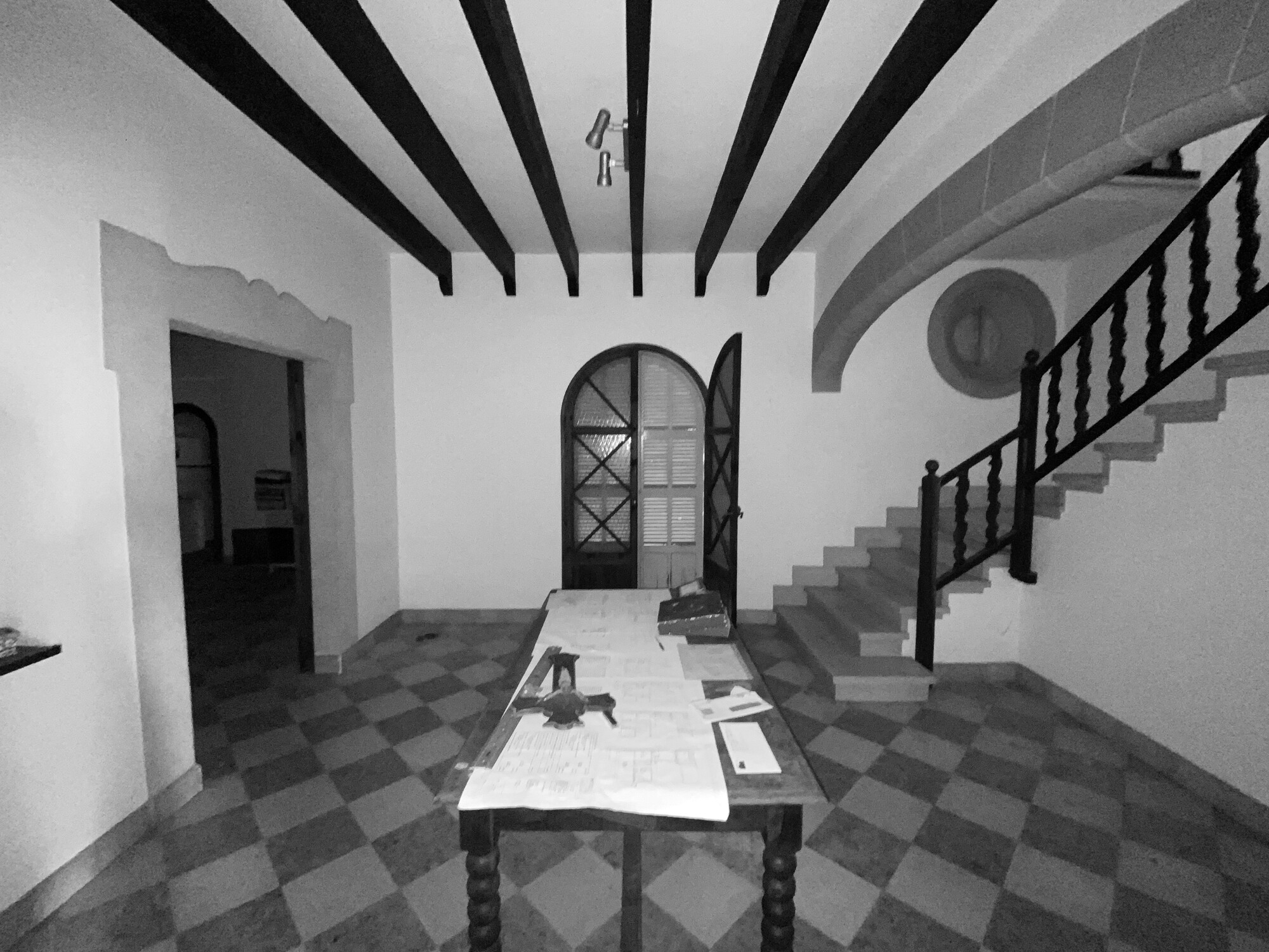 Black and white view of an entrance hall with a checkerboard tile floor, dark ceiling beams, arched door and stone stair, with plans spread on a table