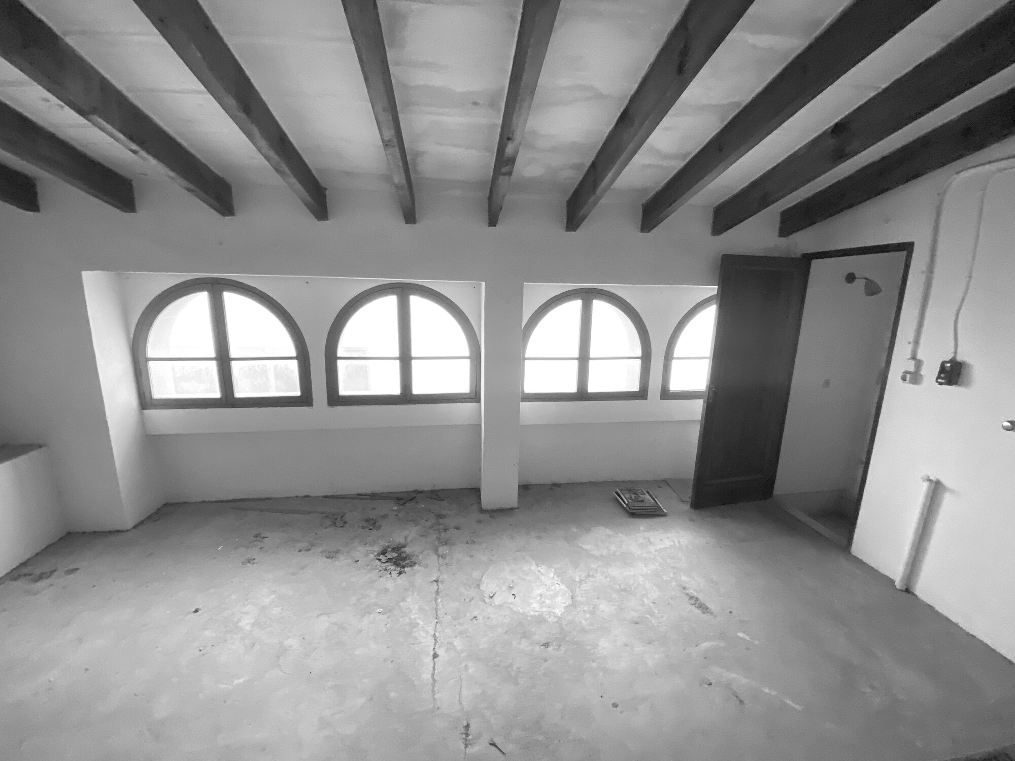 Empty attic room with four arched windows, exposed timber beams and a stained concrete floor in flat daylight