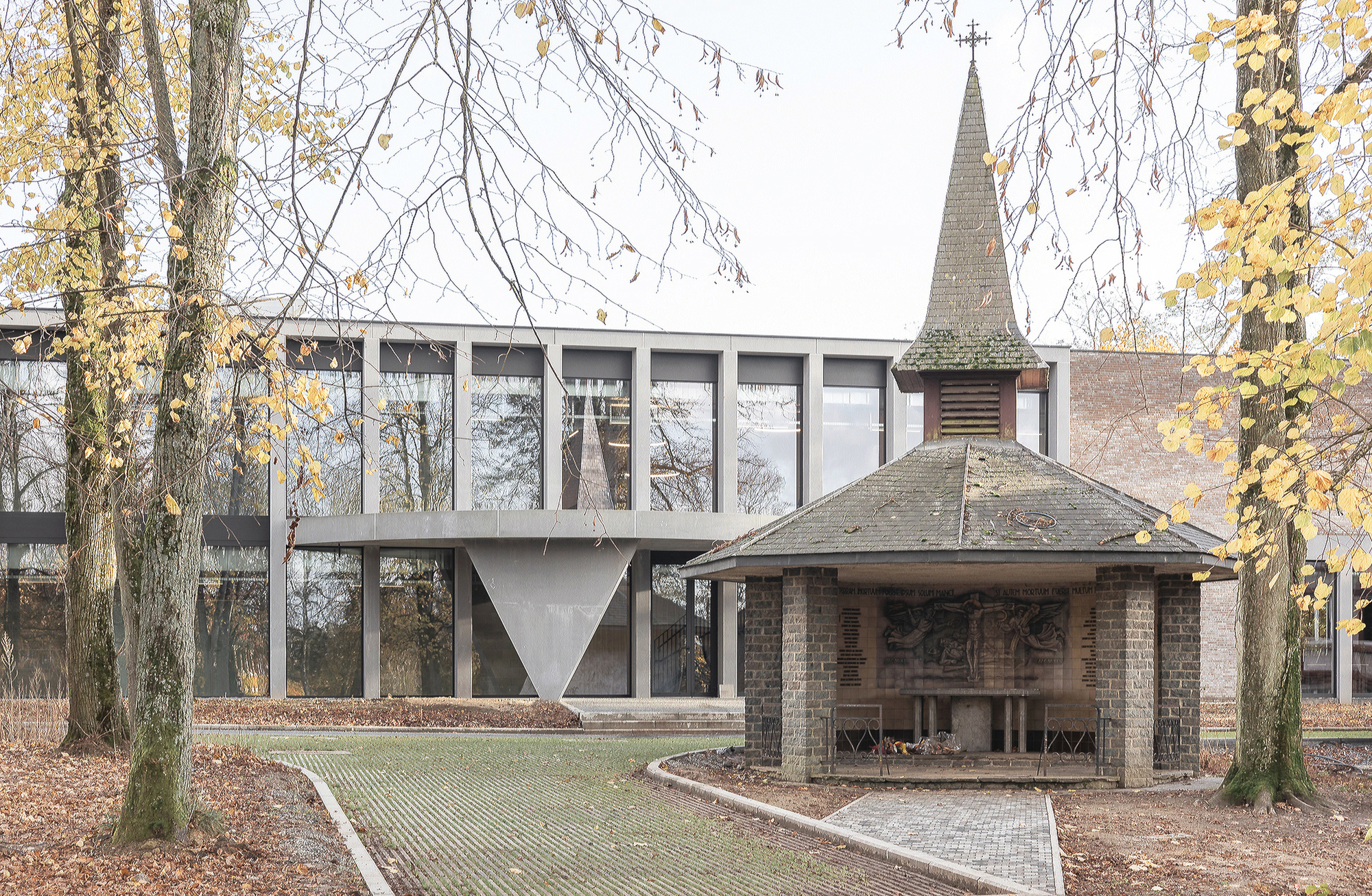 Small brick chapel with a slate spire in front of the glazed concrete-frame wing, framed by yellow-leaved trunks