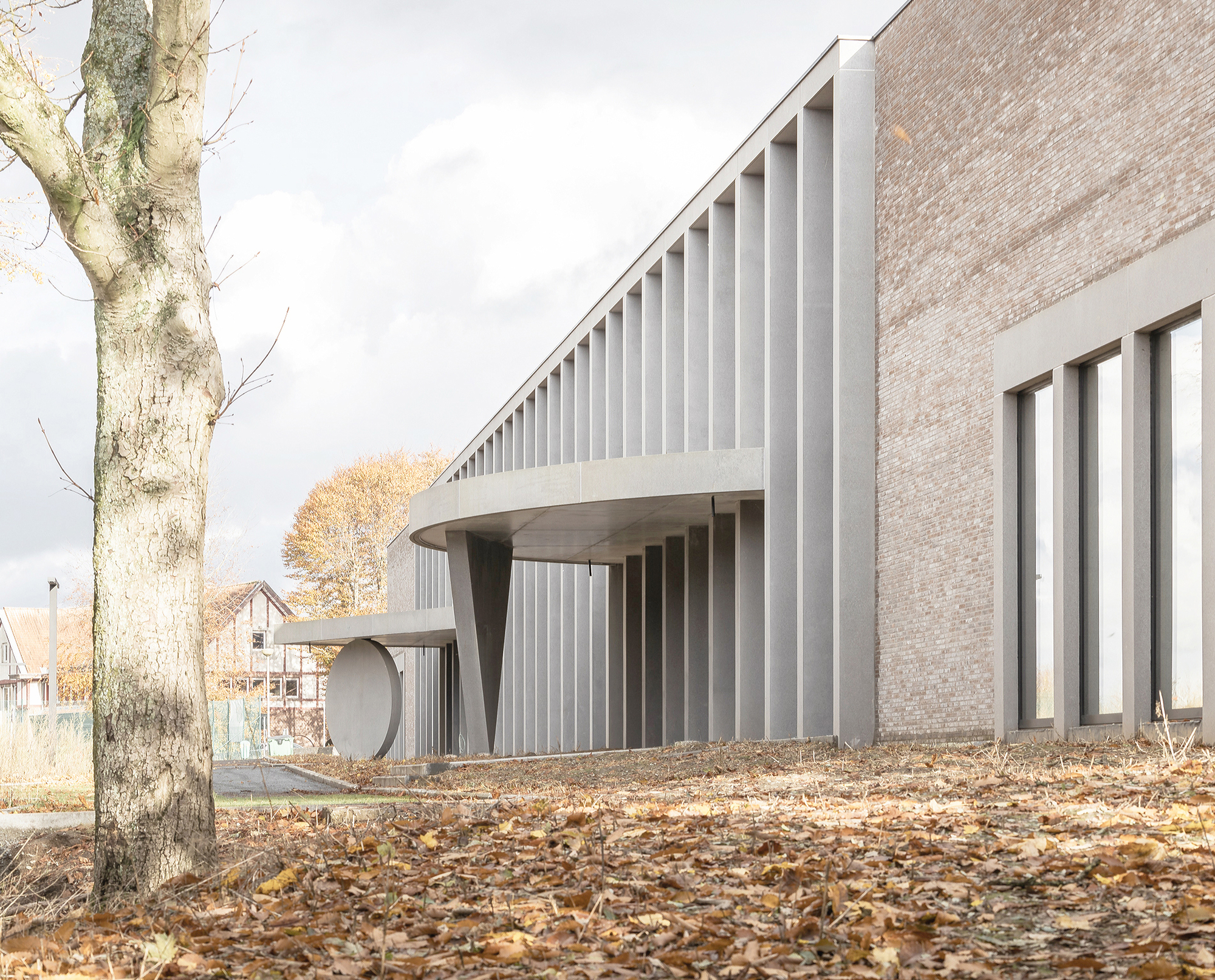 Rhythm of tall concrete fins under a curved canopy beside the brick wall, with a round disc and leaf-covered ground
