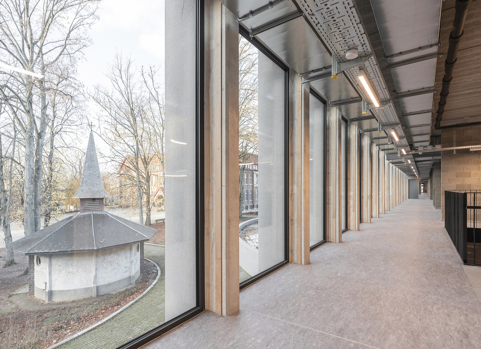 Upper-floor corridor with timber columns and floor-to-ceiling glazing looking onto a small round chapel among bare winter trees