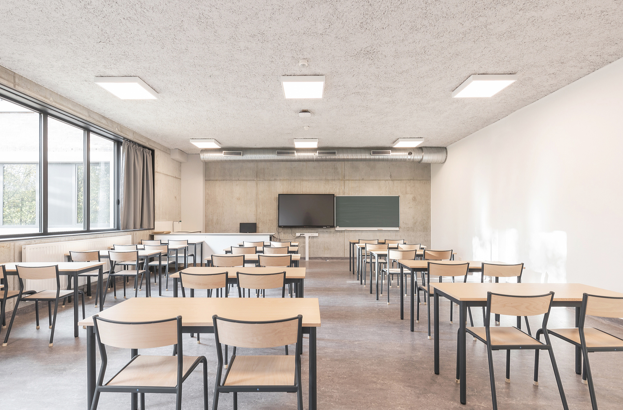 Classroom with light timber chairs and desks, exposed concrete wall, wood-wool ceiling and a window with grey curtain in daylight