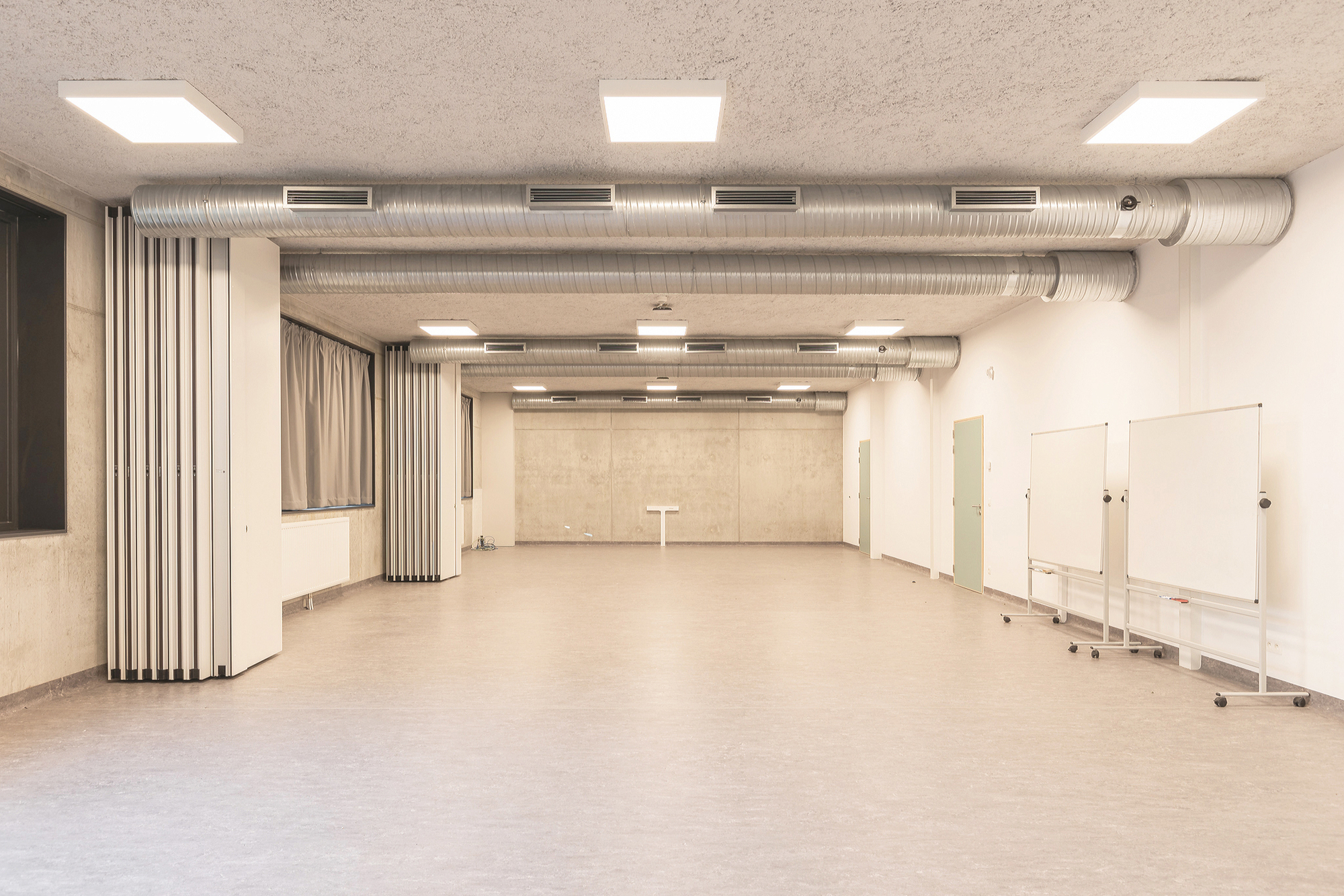 Open multipurpose room with folding partition walls, exposed spiral ducts, square ceiling lights and two mobile whiteboards on the right