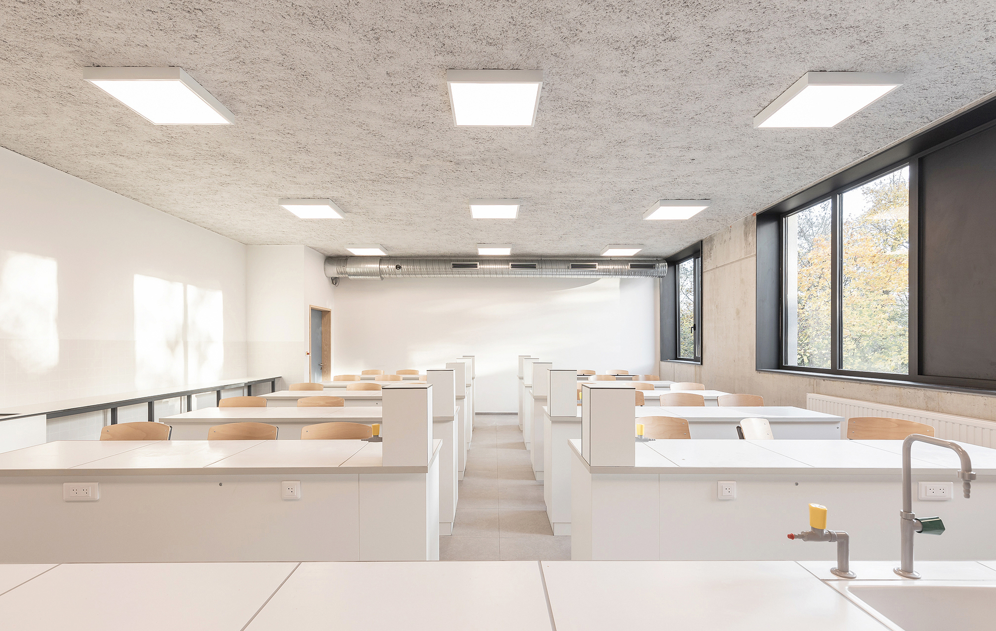 Science classroom with white lab benches, wooden stools, square ceiling lights and a tall window onto autumn trees