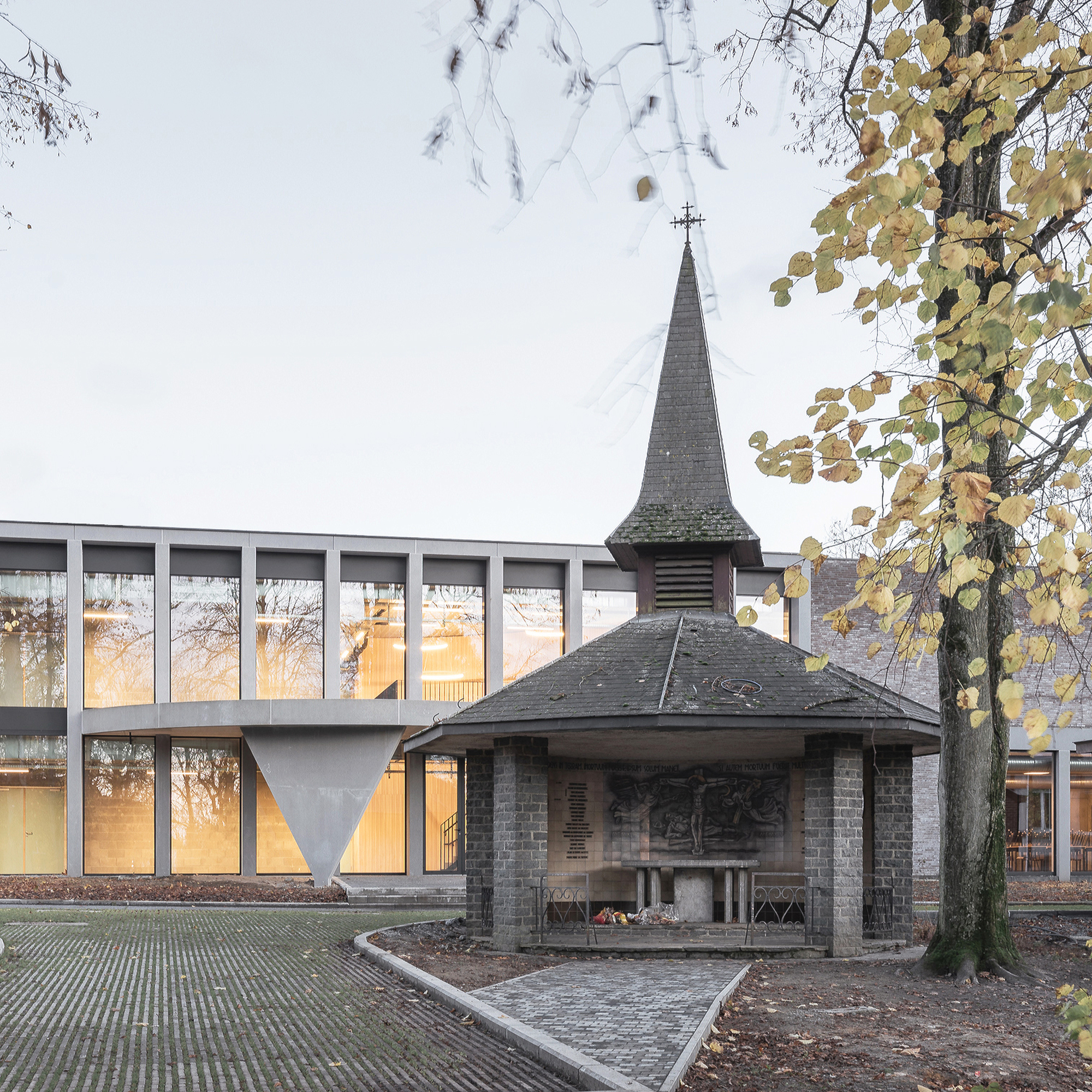 Small brick chapel with slate spire in front of a glazed two-storey facade, framed by a yellowing tree