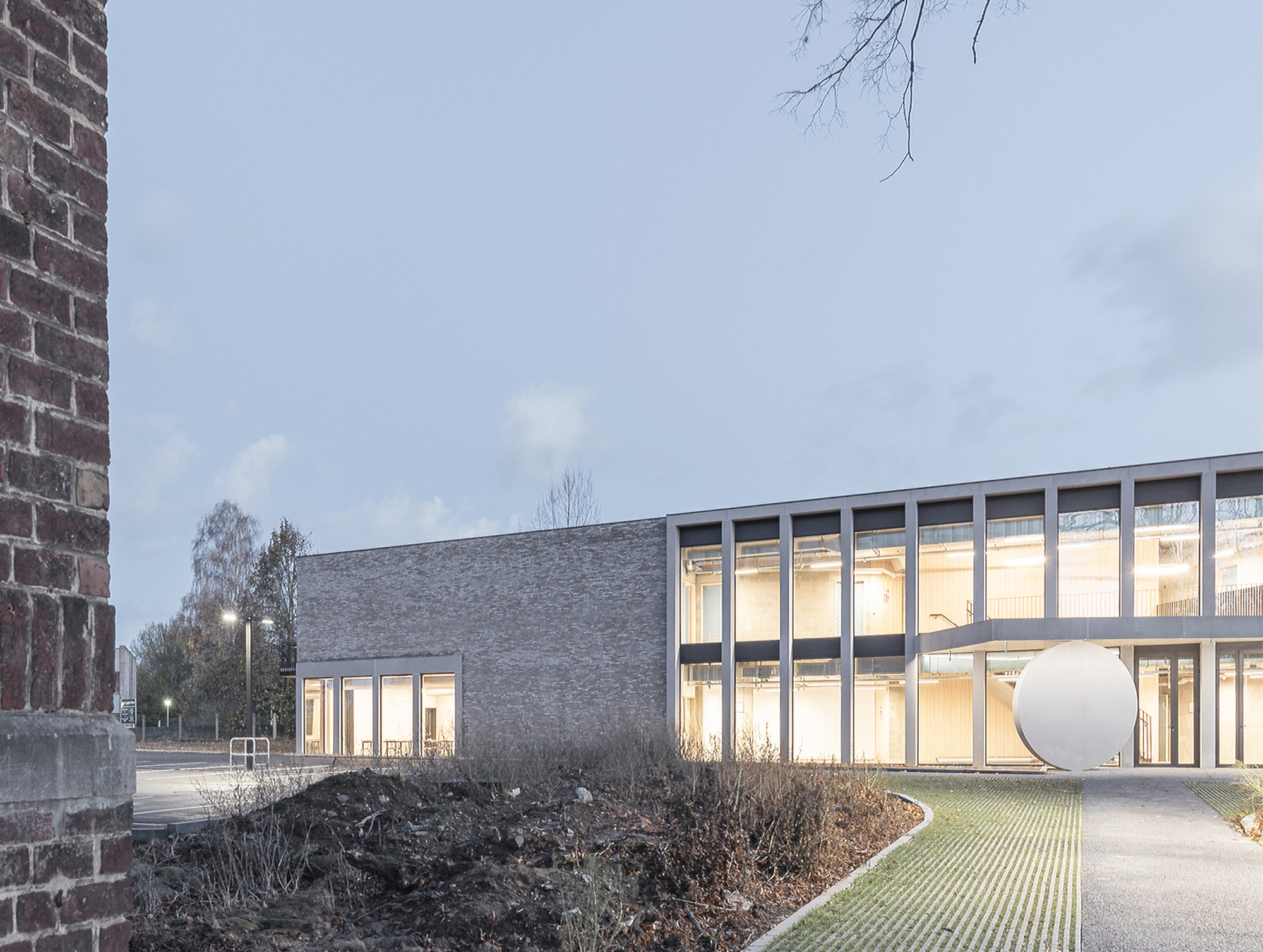 Glazed gridded facade beside a grey brick volume, with a round white disc sculpture at dusk