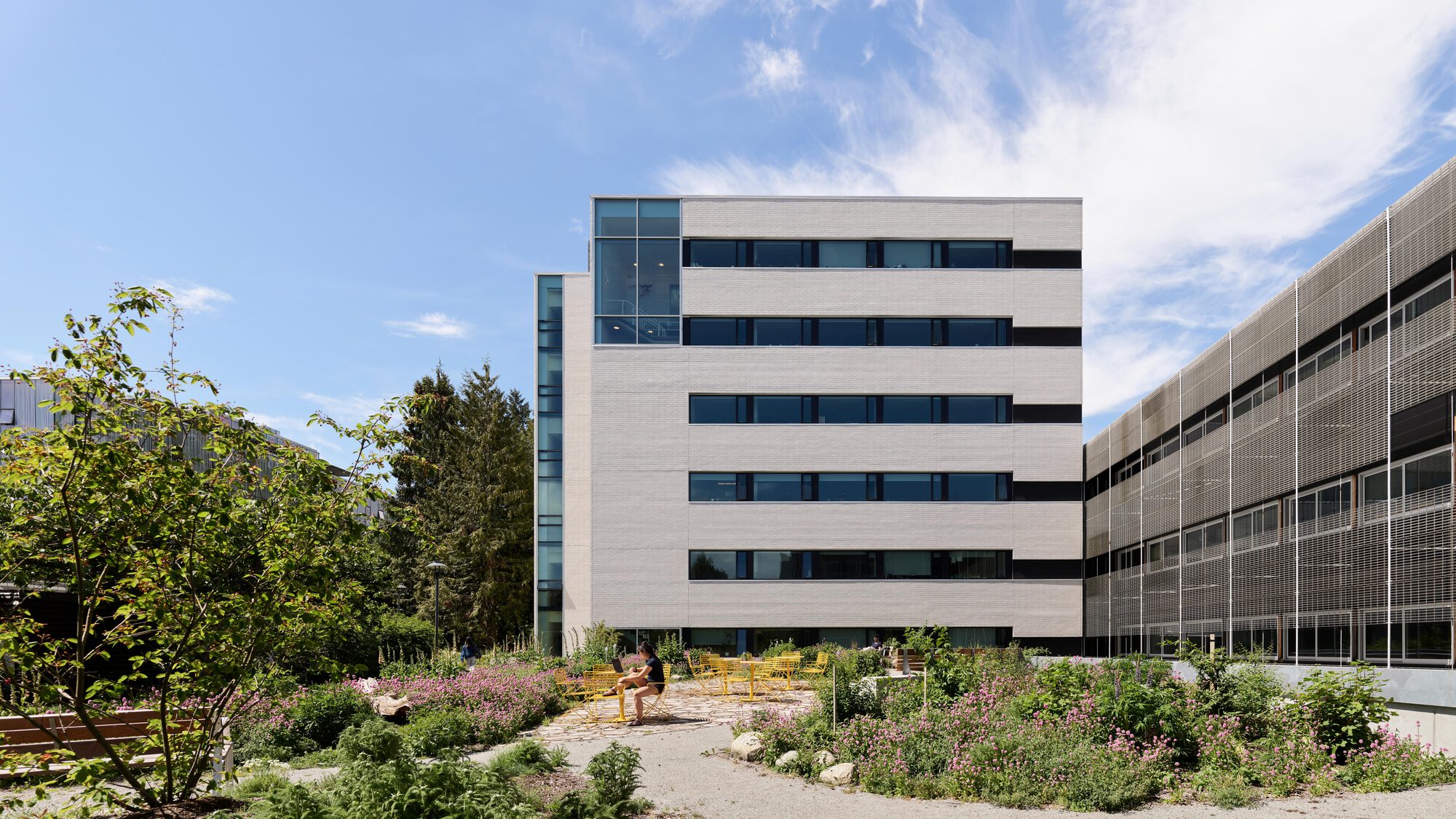 Pale banded masonry block with dark window strips above a courtyard garden of flowering perennials, a person seated on a yellow chair
