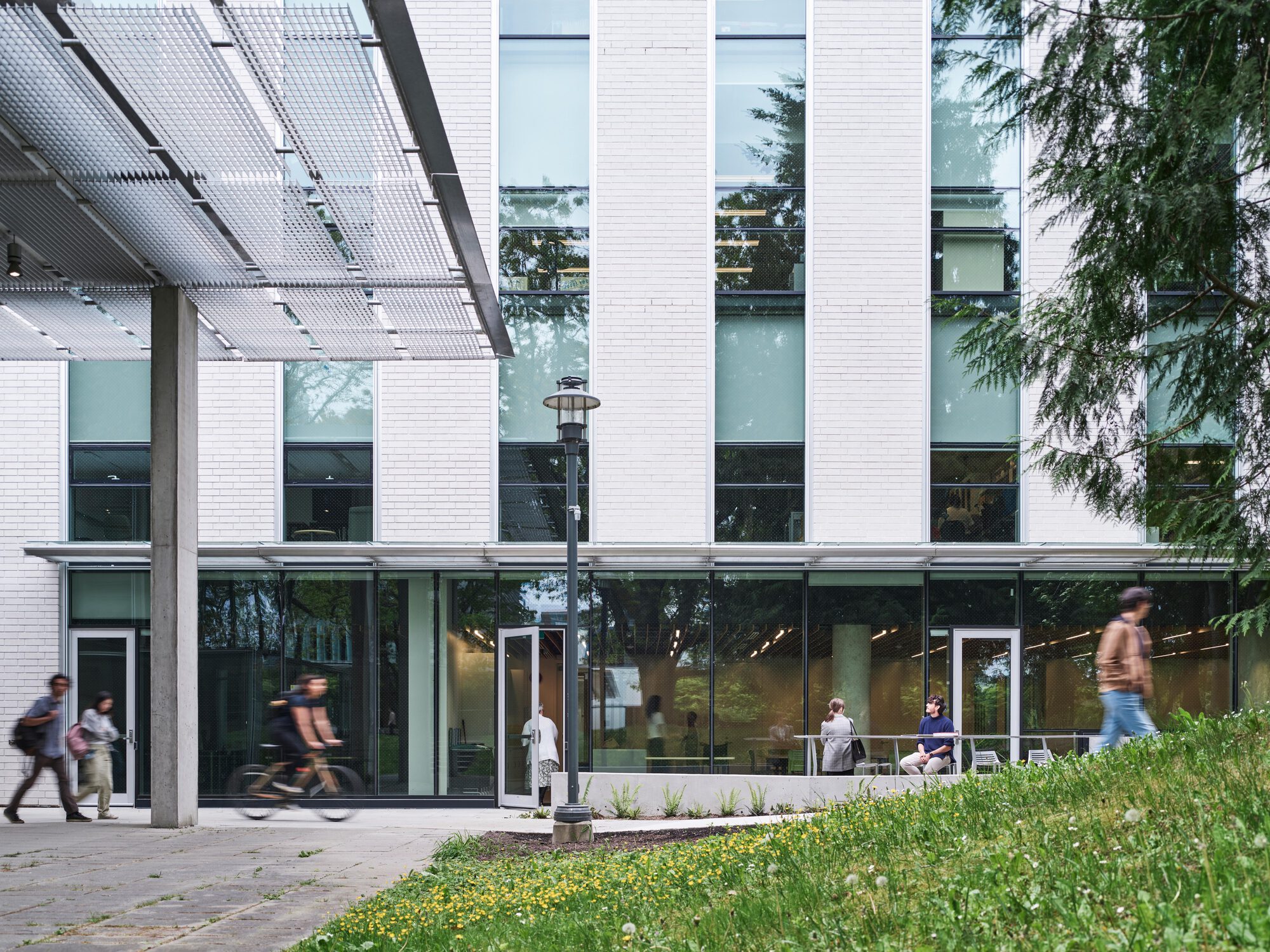 Concrete column and mesh canopy beside the glazed ground floor, with a cyclist and walkers crossing in front of a grassy bank