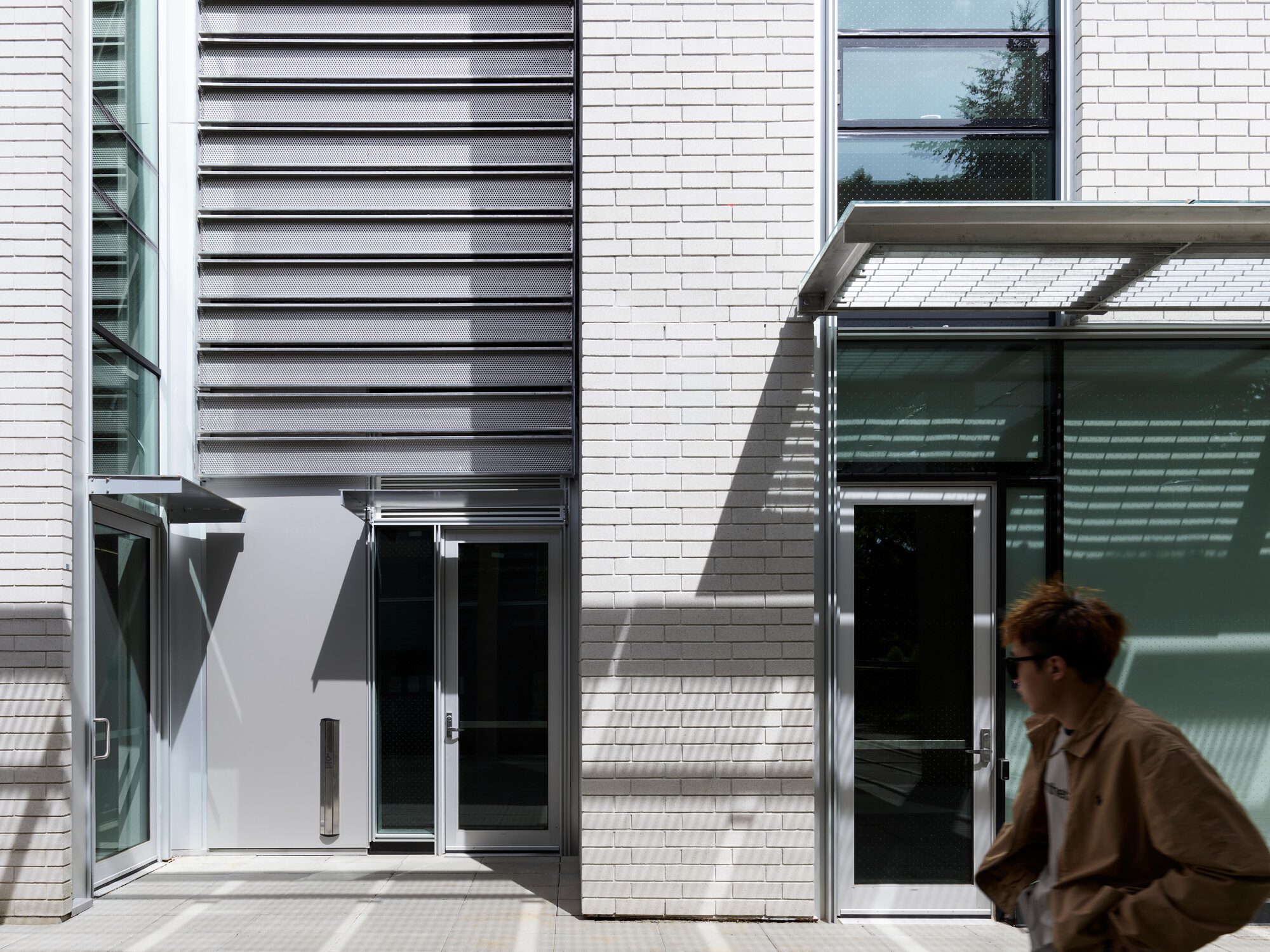 Ground-floor entrances with louvered metal panel, glass doors and a mesh canopy, as a passerby in a tan jacket walks past