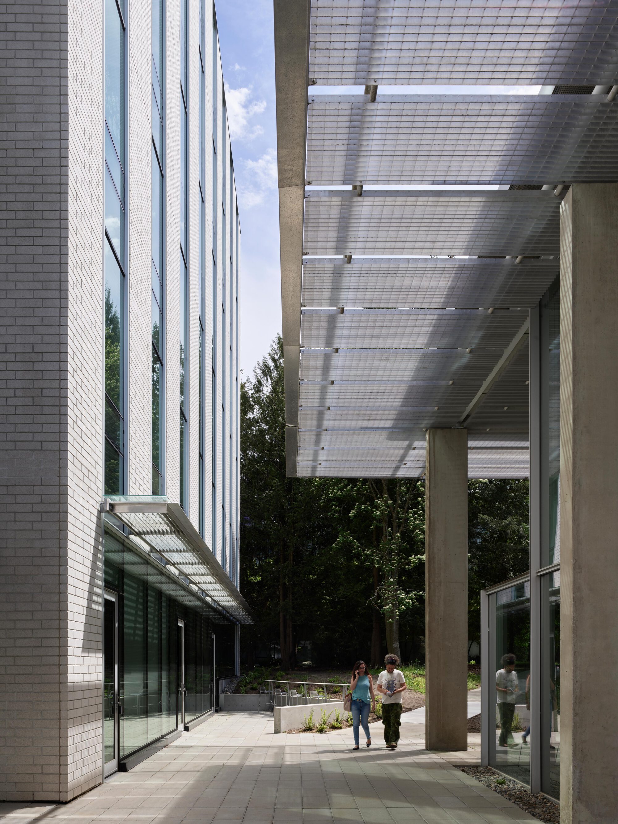 Walkway between a brick and glass facade and a concrete-columned mesh canopy, with two people strolling toward trees under blue sky
