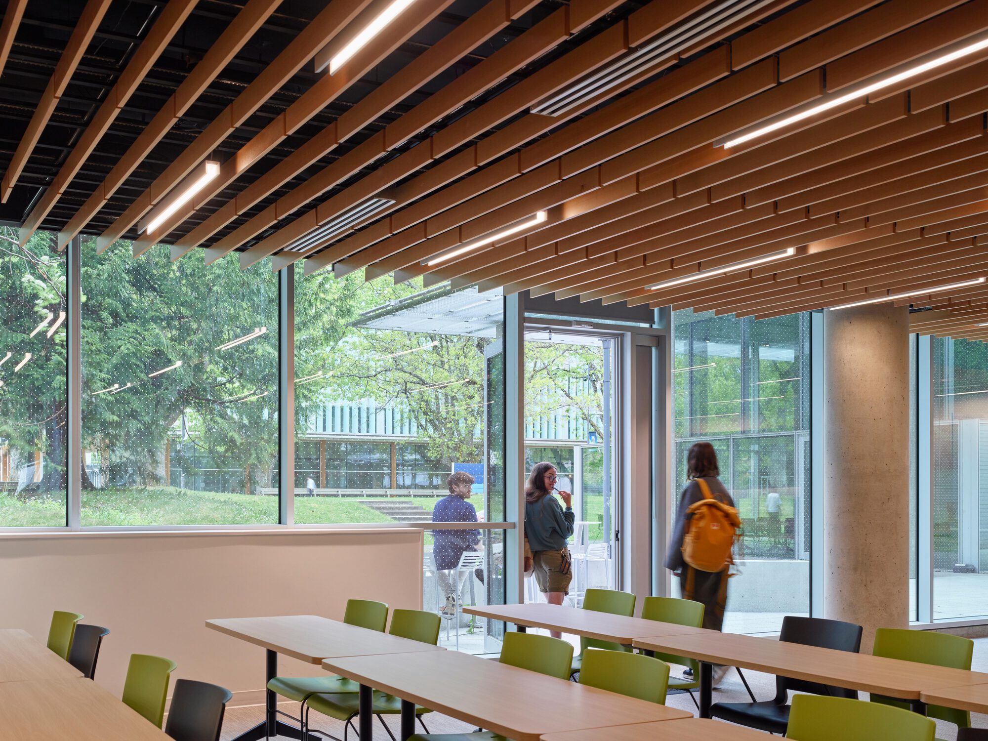 Classroom with timber slat ceiling and linear lights, green chairs and long tables, where students stand by an open glass door to the lawn