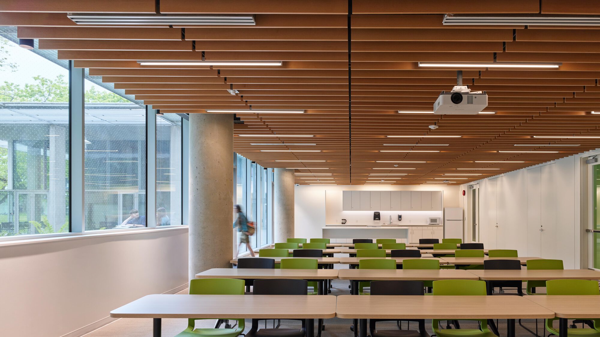 Classroom with green chairs and long tables beneath a timber slat ceiling, a student walking past the columns and glazing in daylight