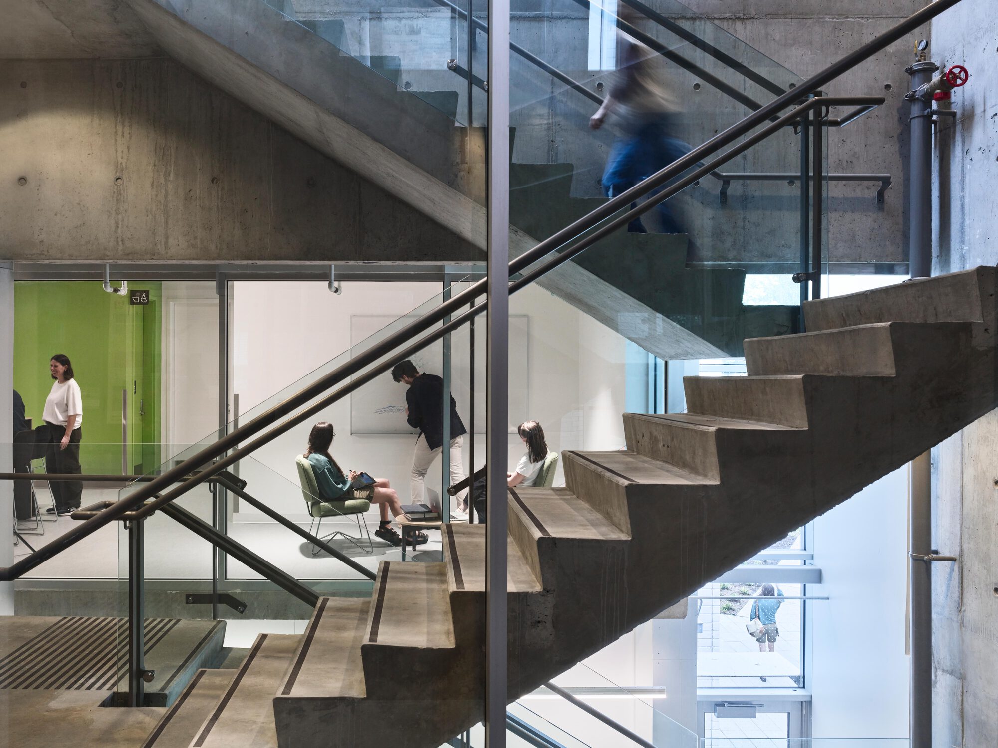 Exposed concrete stair with glass balustrades, people meeting in a glazed room beyond and a blurred figure climbing above