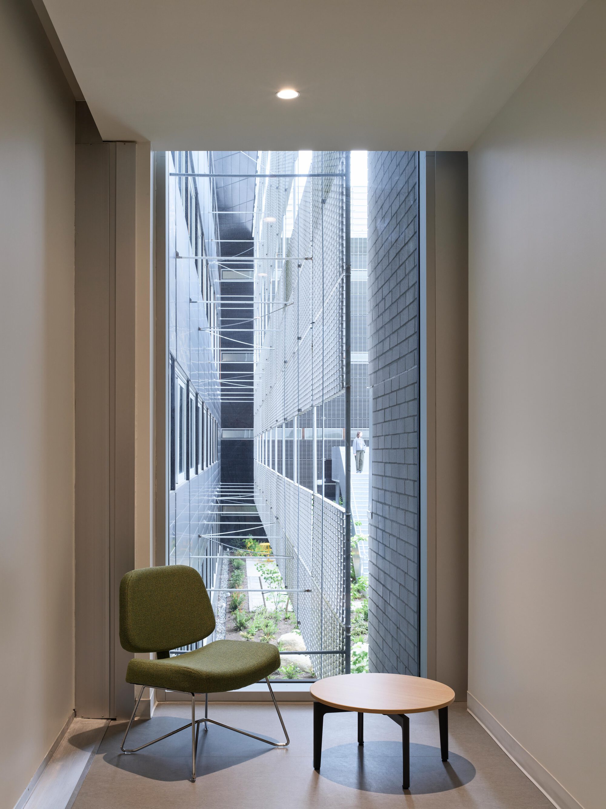Corridor nook with a green lounge chair and round side table facing a tall window onto a narrow planted courtyard