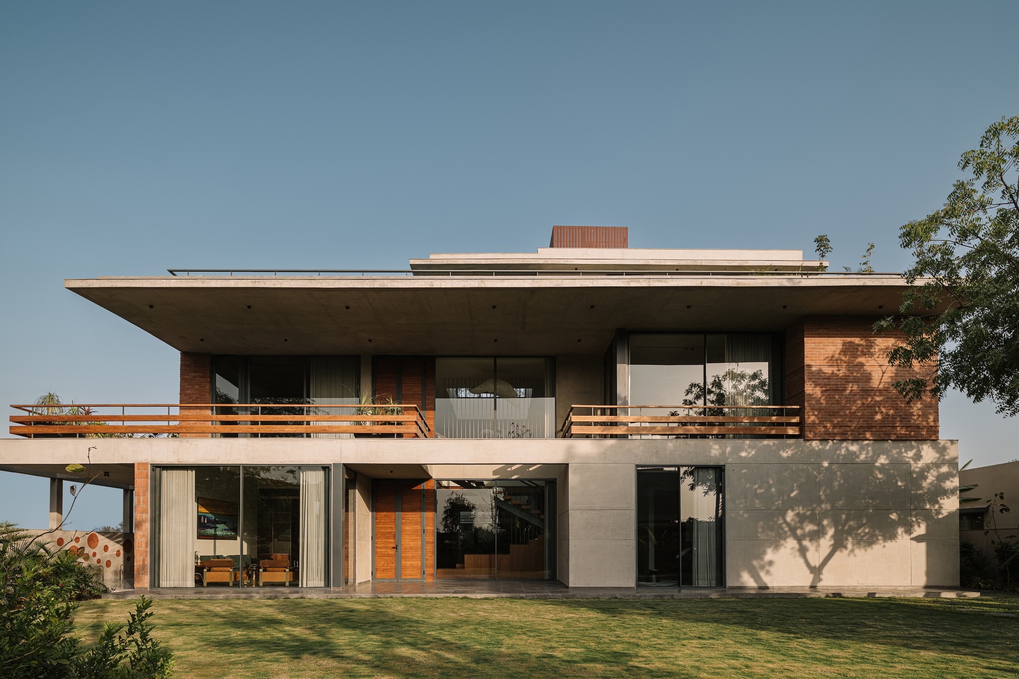 Front facade of the two-storey house with a deep concrete roof slab, timber balcony rails and brick panels, a tree shadow across the lawn